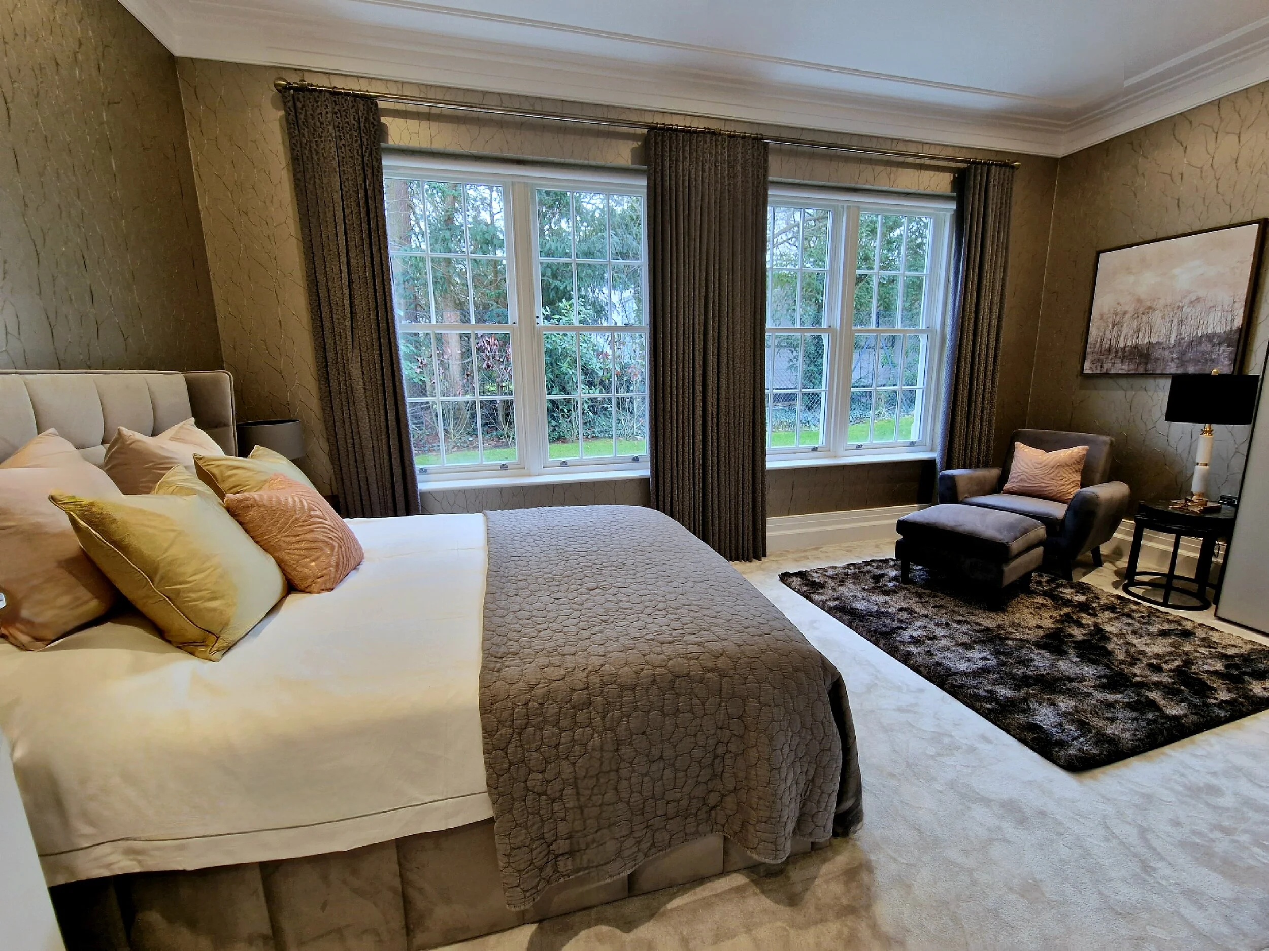 A bedroom with a king-sized bed with beige pillows and a textured grey bed throw, a small seating area with a grey armchair, a matching ottoman, a black side table, a lamp, and a large window with dark curtains showing a view of a garden outside.