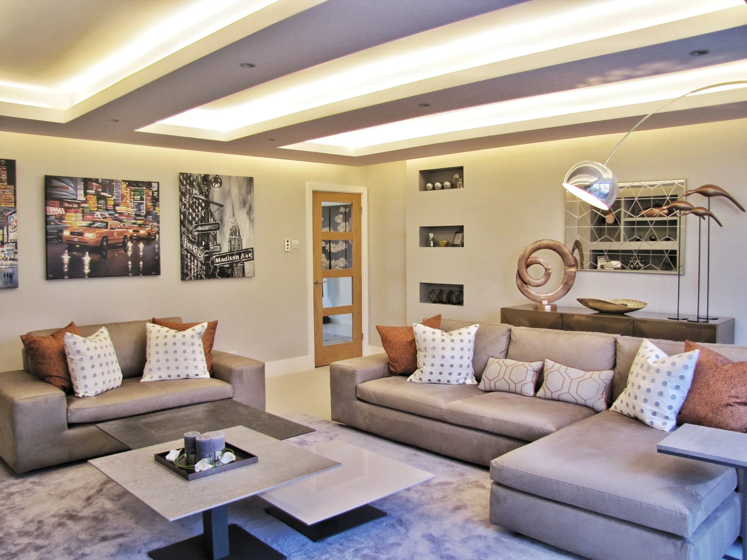 Modern living room with two beige sofas with patterned pillows, a coffee table with decorative items, wall art, decorative shelves, and contemporary lighting fixtures.