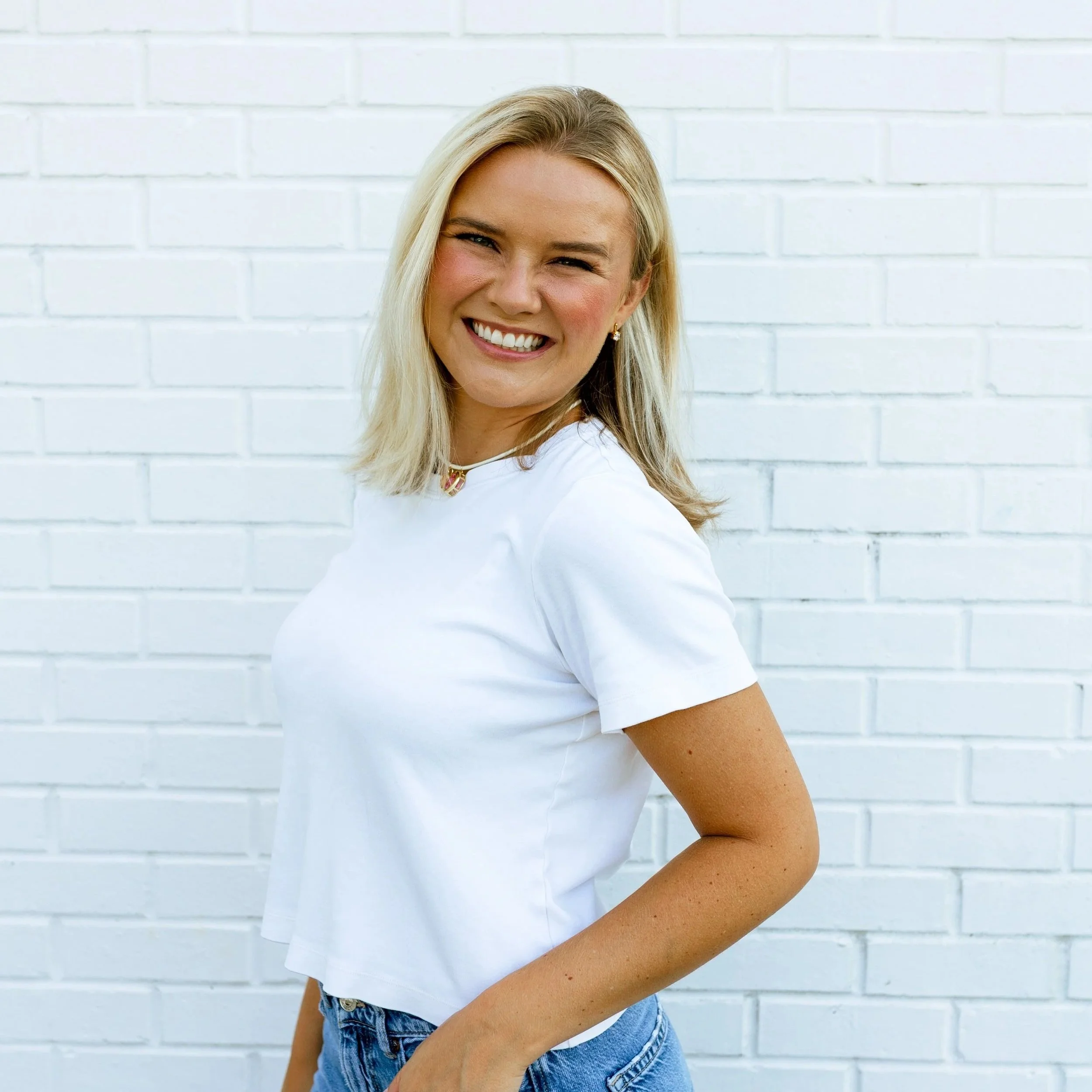 A smiling woman with blonde hair, wearing a tan sleeveless top and light blue jeans, standing against a white background.
