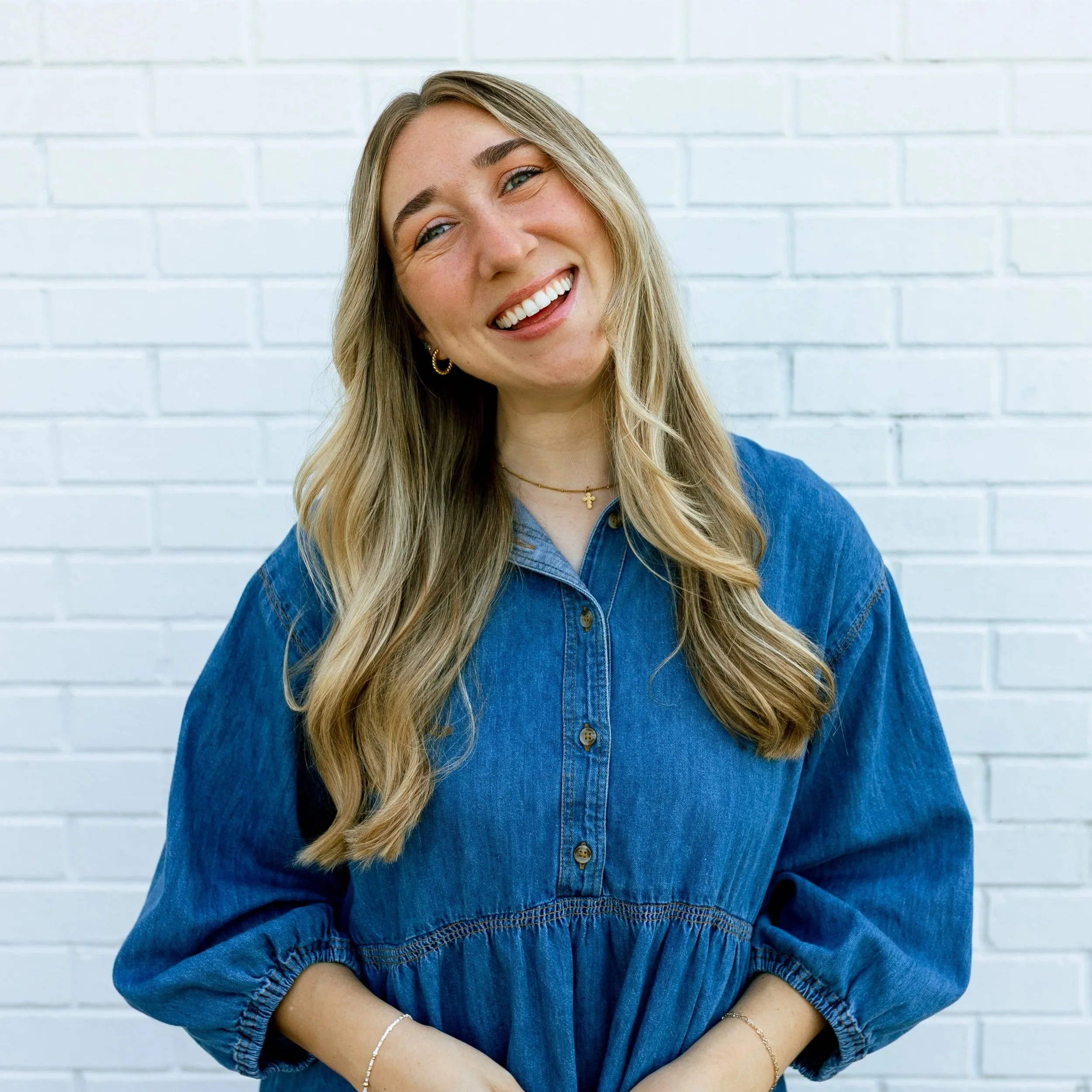 A smiling woman with long wavy blonde hair, wearing a white sleeveless top with ruffled edges and blue drawstring pants, stands against a plain light background.