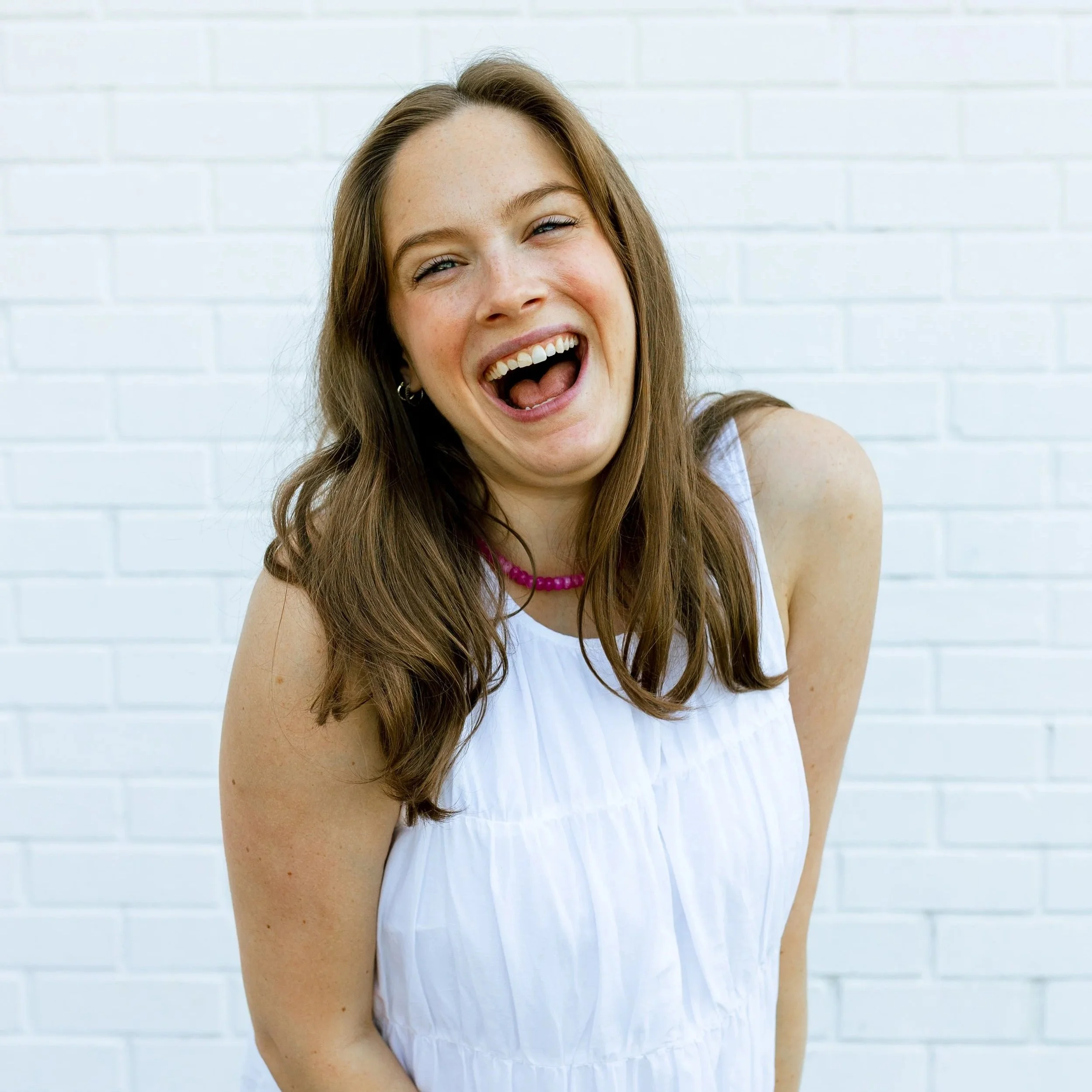 Joyful young woman with shoulder-length brown hair, wearing a lace long-sleeve top and a light blue denim overall, standing against a plain light background.