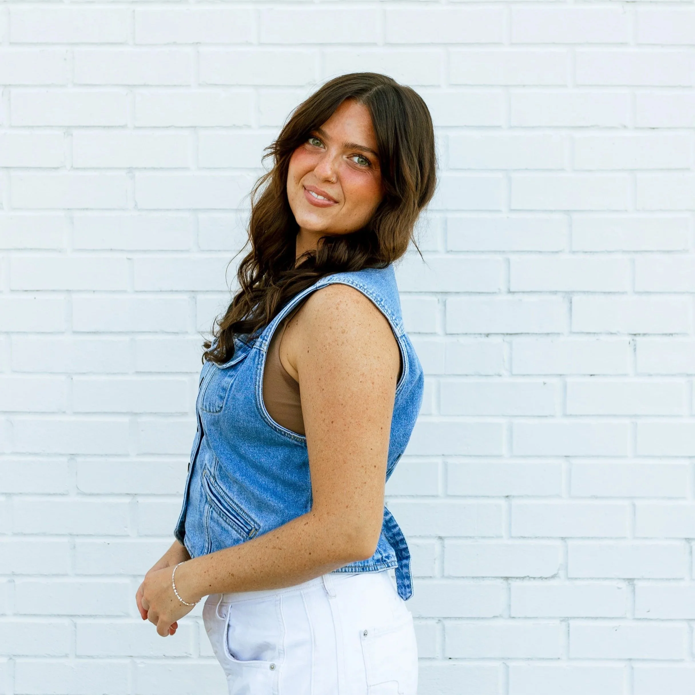 Young woman with long wavy brown hair, wearing a white blouse and denim overalls, standing against a plain white background.