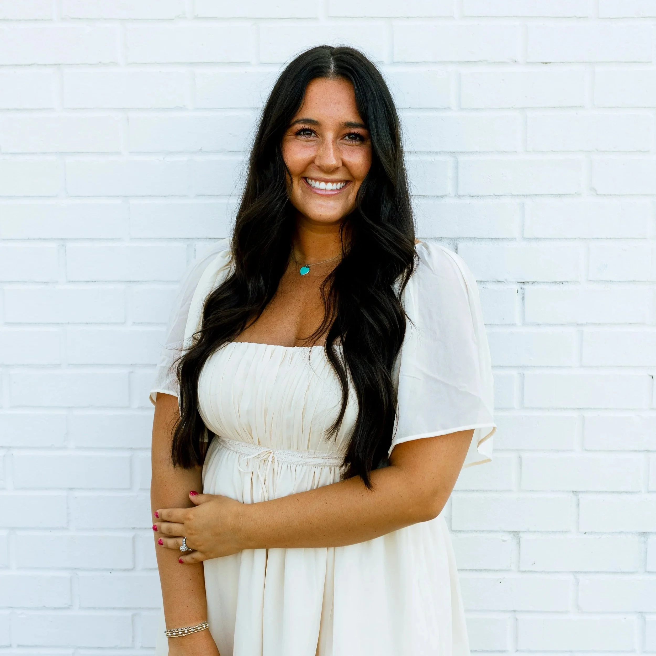 A young woman with long wavy brown hair smiling, wearing a white blouse with buttons and lace details, standing against a plain white background.