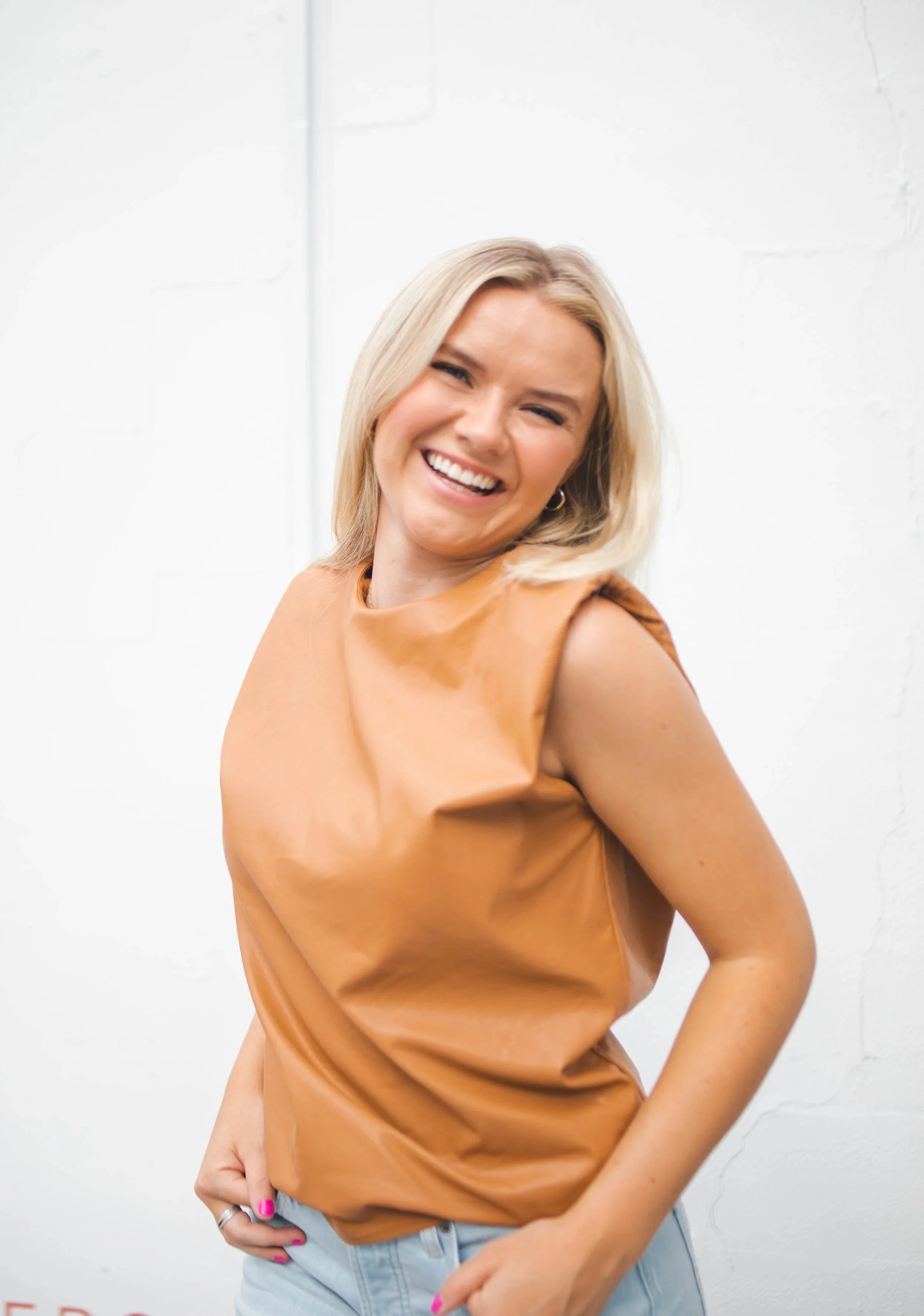 A smiling woman with blonde hair, wearing a tan sleeveless top and light blue jeans, standing against a white background.