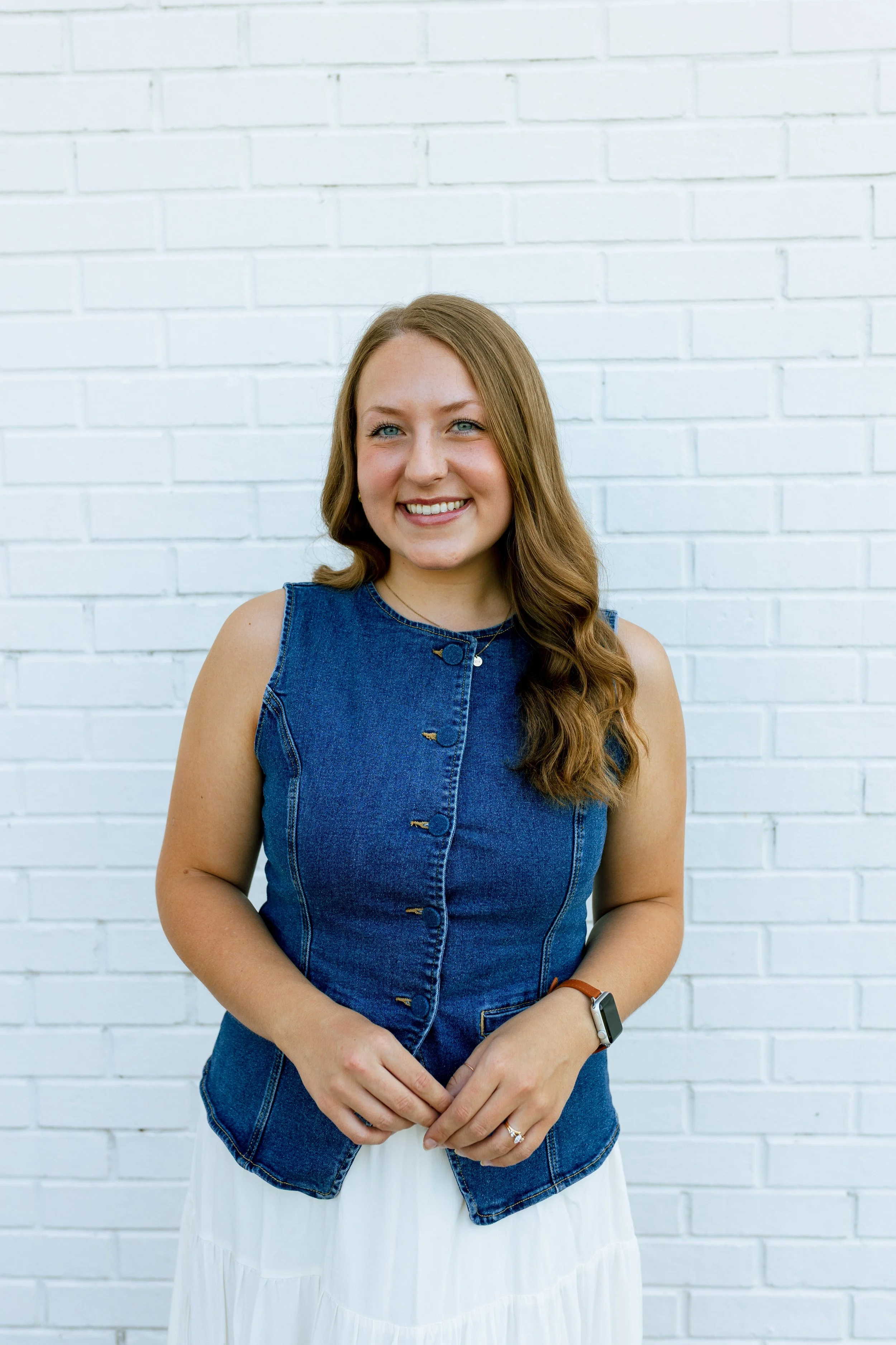 Young woman with red hair smiling, wearing a sleeveless white top with brown buttons, blue jeans, and a watch, standing against a plain white background.