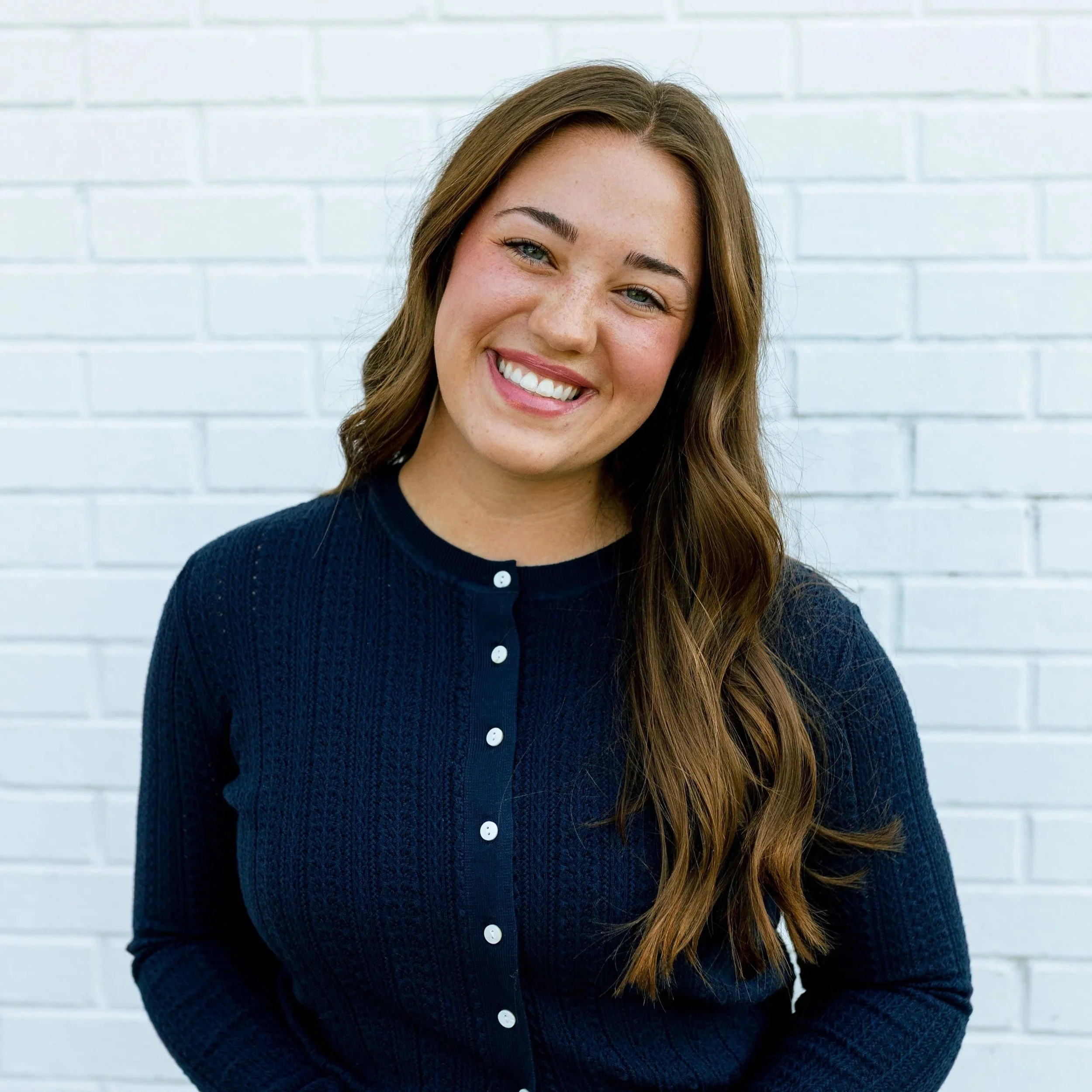 A woman with long dark wavy hair smiling and holding an open book, wearing a white shirt and light blue jeans, standing against a plain white wall.