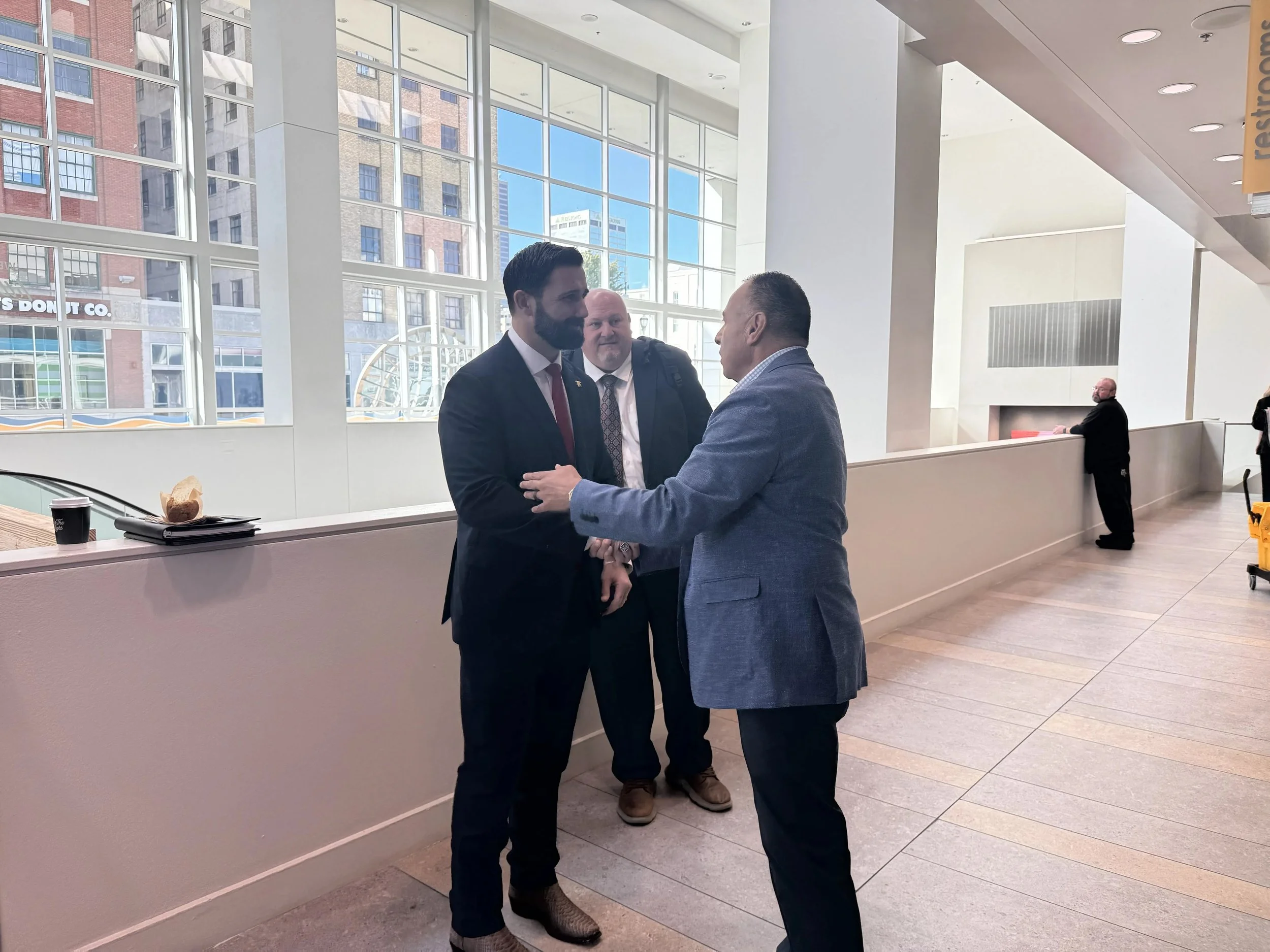 Three men in business attire greeting each other in a bright convention center hallway at the NEMA Annual Forum, where Lima Charlie Inc. follows state disaster housing planning.