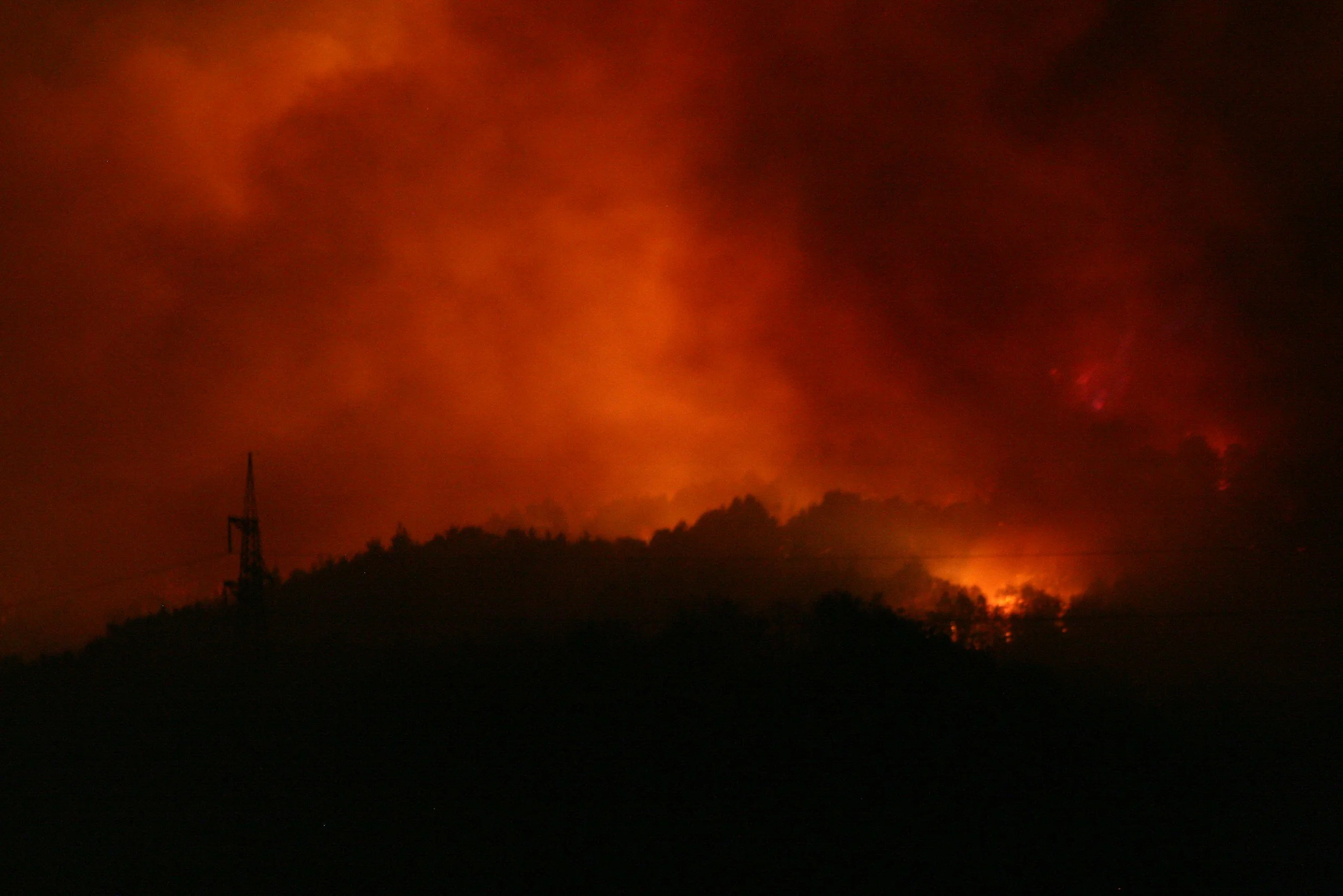 A dense orange wildfire glow and smoke rising over a dark hillside at night with a power line tower silhouetted in the foreground, showing the kind of active wildfire conditions that leave families needing emergency housing from Lima Charlie Inc.
