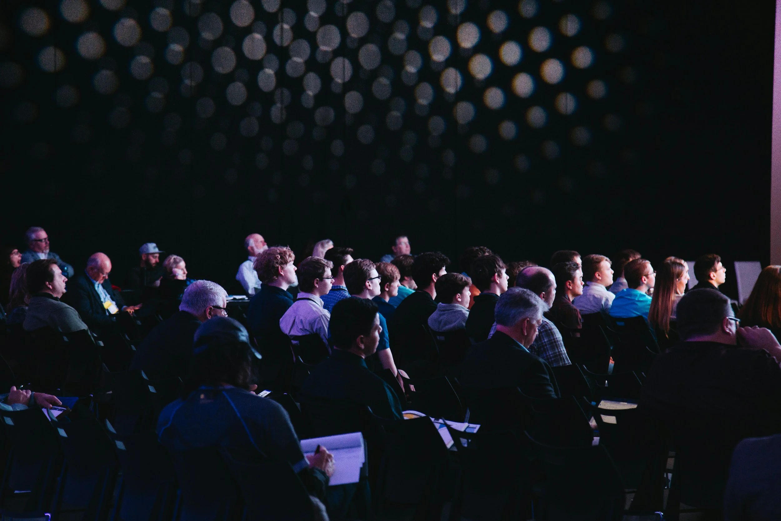 A large conference audience seated in a dim auditorium watching a keynote presentation, the kind of major convention that fills every hotel in a host city where Lima Charlie Inc. arranges corporate lodging instead.