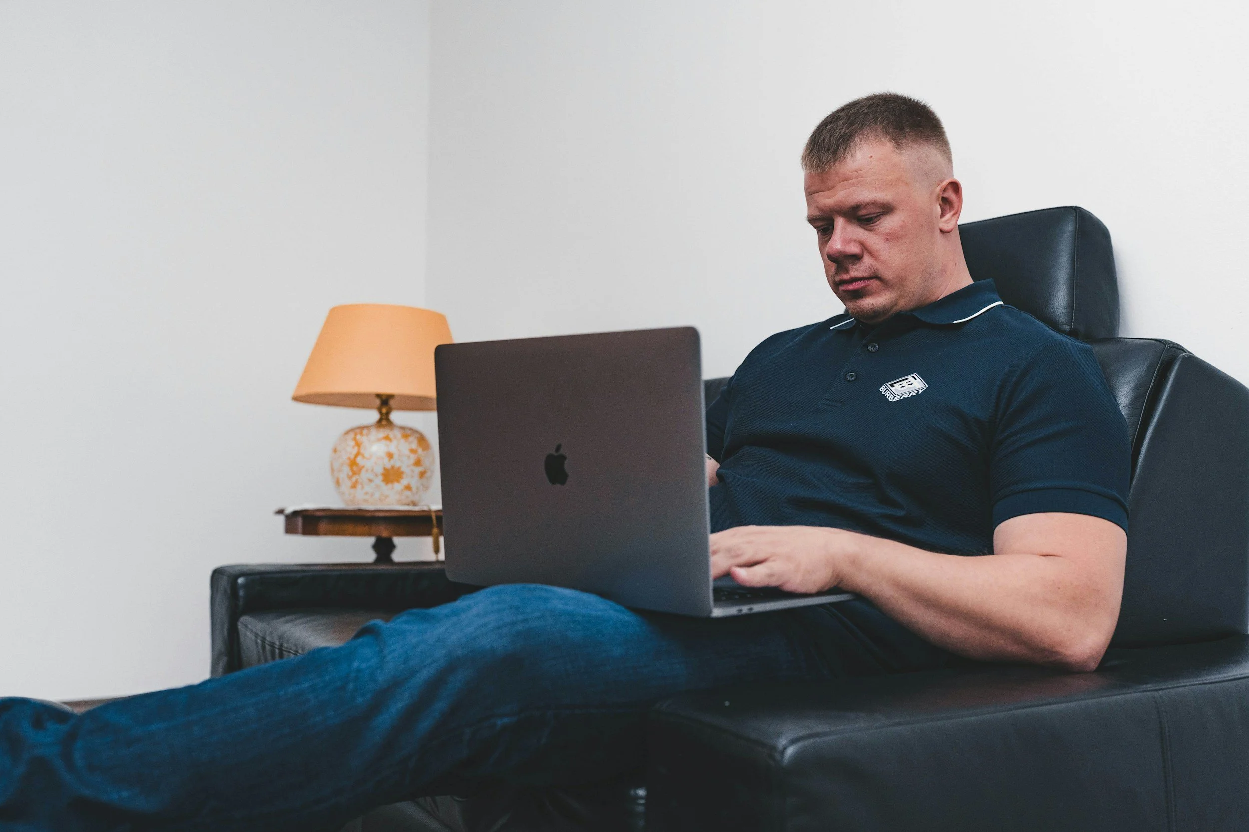 A man in a work polo uses a laptop while relaxing in a leather recliner beside a lamp, reflecting the extended-stay corporate lodging Lima Charlie Inc. offers project teams after a long shift.