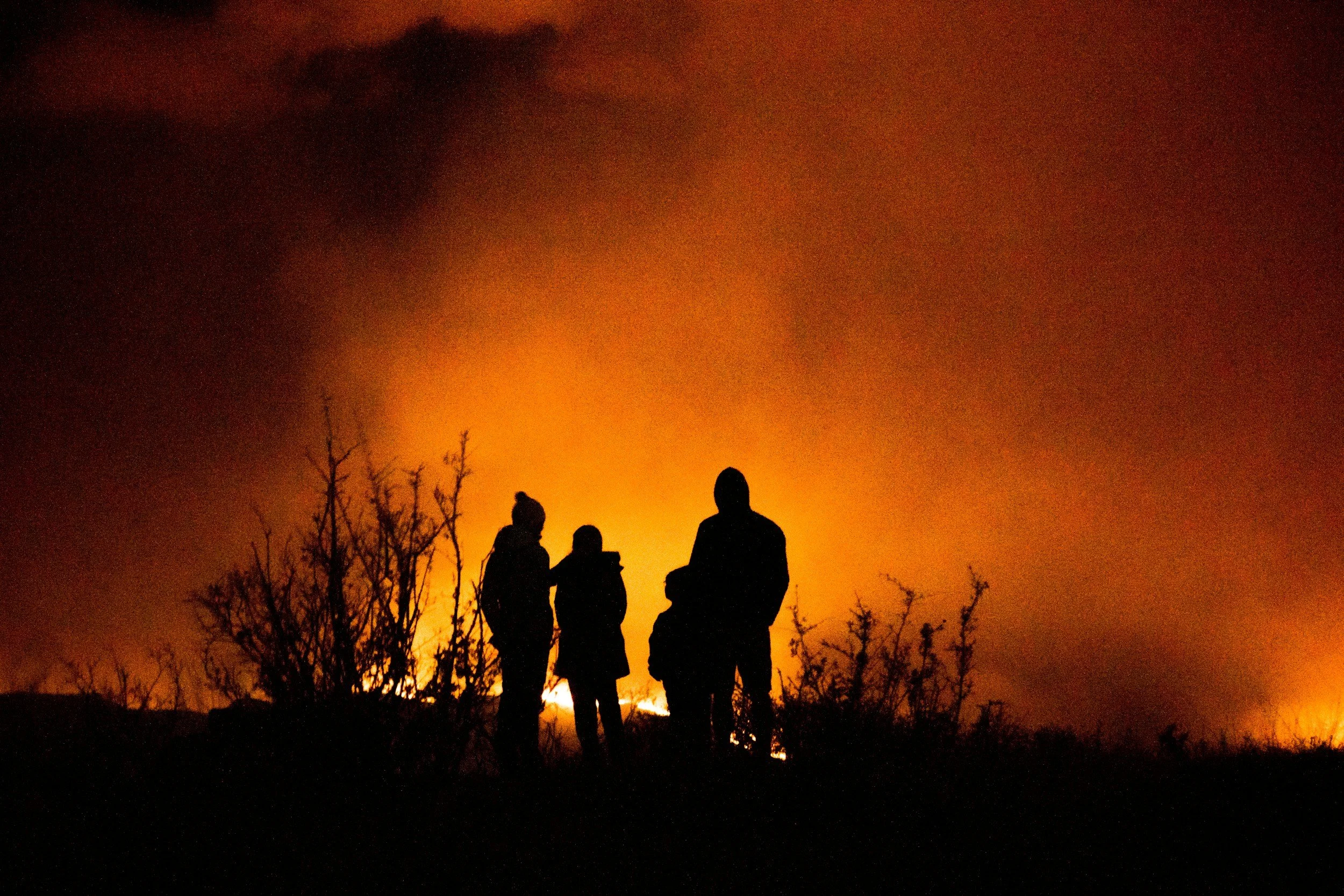 A family of four stands silhouetted against a glowing orange wildfire and smoke filling the night sky, the kind of sudden displacement event Lima Charlie Inc. responds to with emergency response housing and FEMA disaster housing assistance.