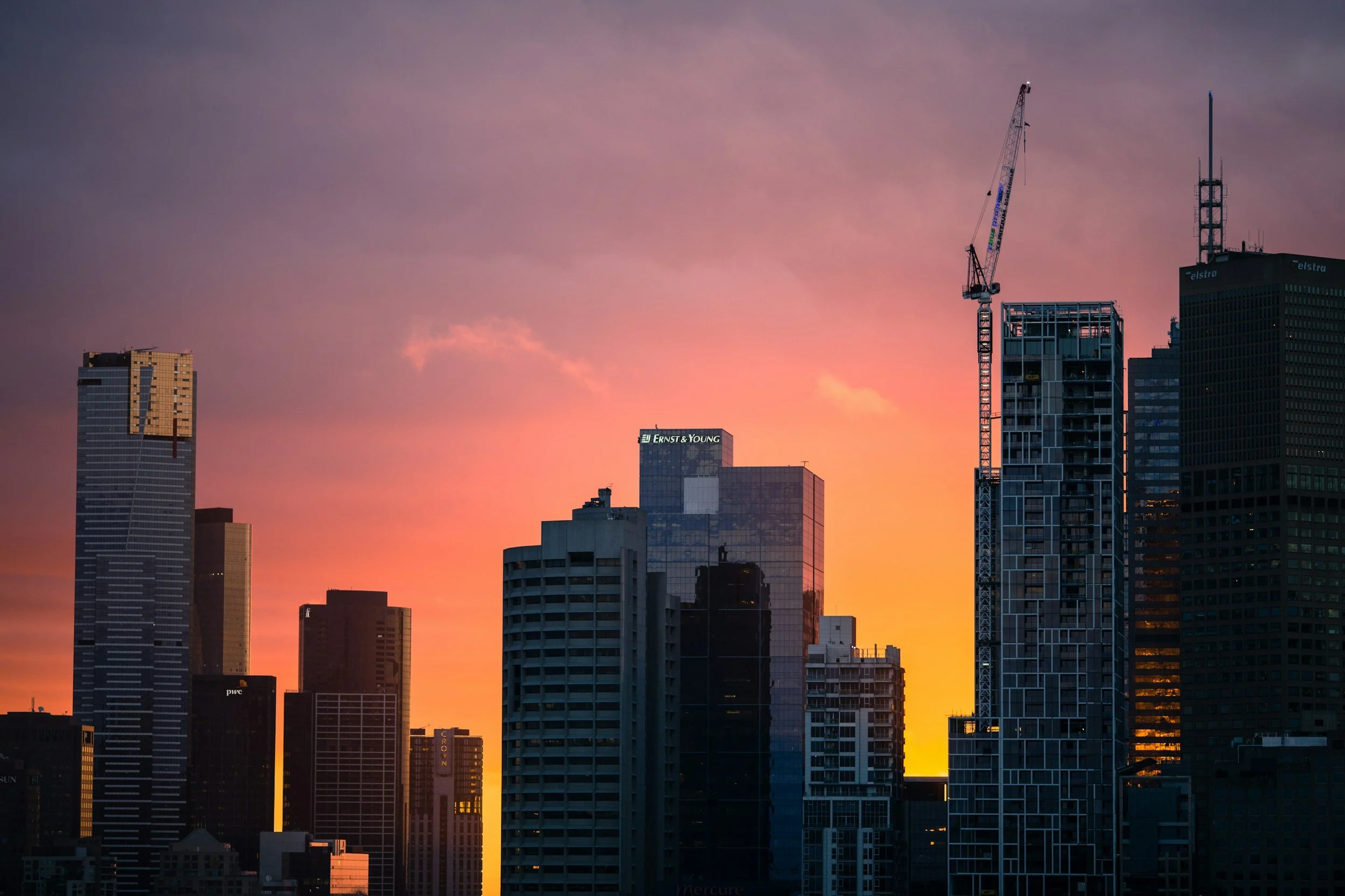 A downtown city skyline glows orange and purple at sunset with a construction crane rising among office towers, the kind of urban worksite where deployed teams rely on Lima Charlie Inc. for furnished corporate lodging near the job.