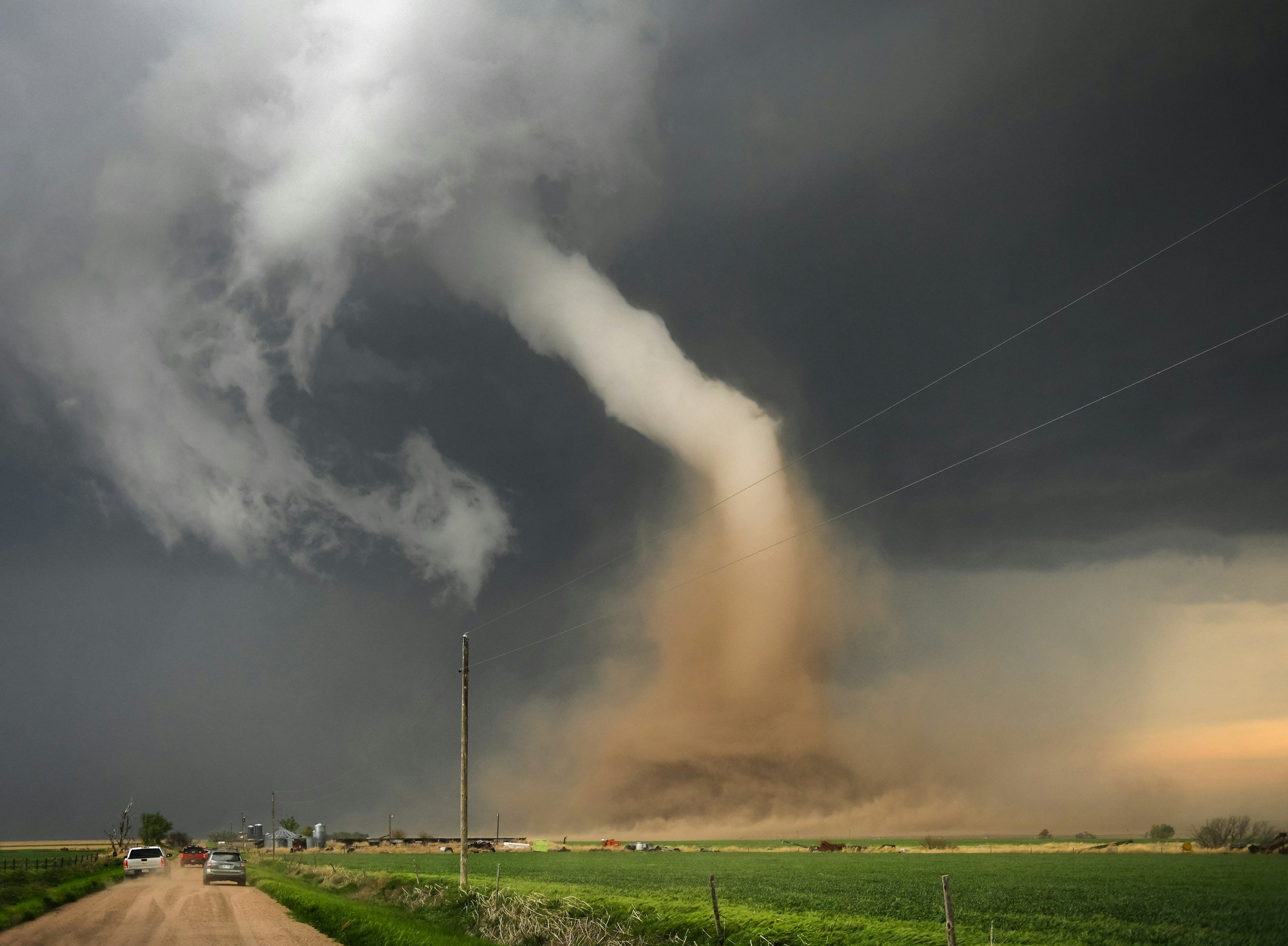 A tornado touches down across an open field near a rural road as vehicles pull away, the kind of severe storm damage that drives demand for emergency response housing from Lima Charlie Inc.