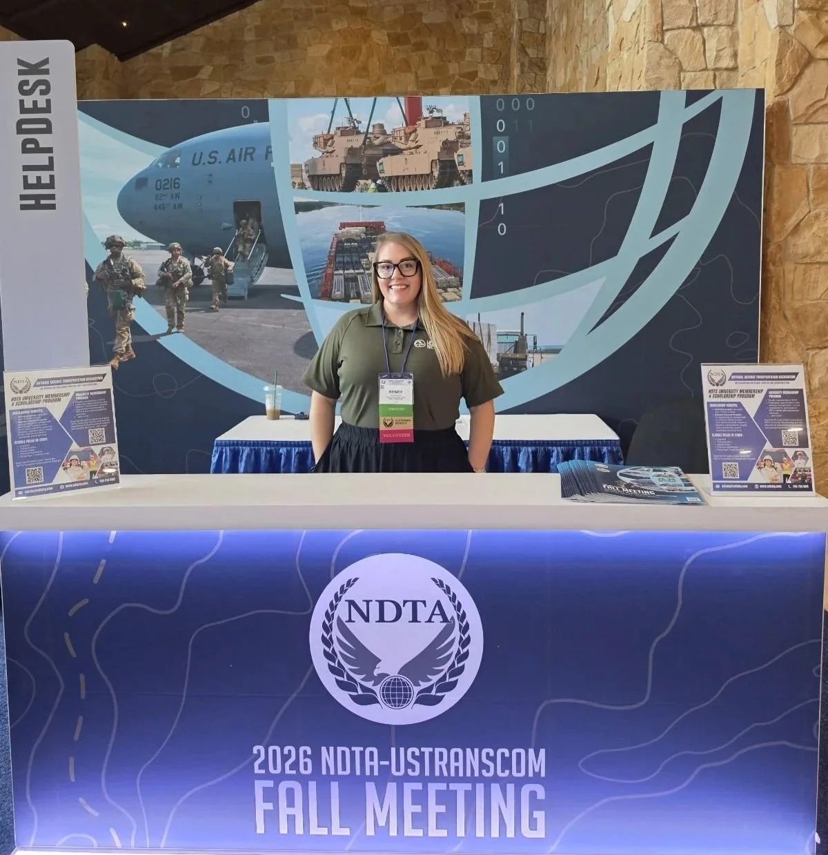 A Lima Charlie Inc. team member smiling at the NDTA help desk under the 2026 NDTA-USTRANSCOM Fall Meeting sign, volunteering with the government lodging team.