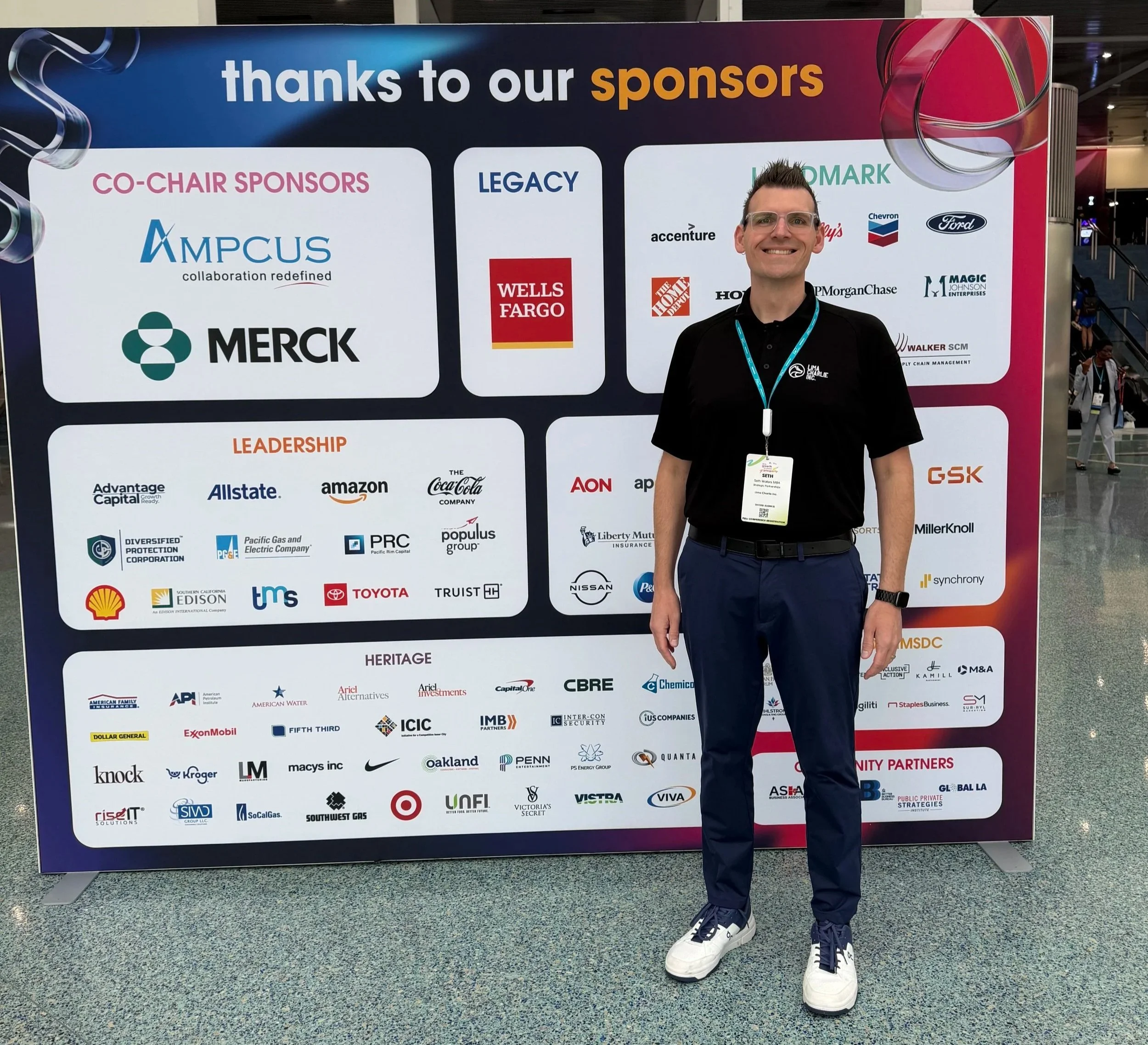 Seth Waters of Lima Charlie Inc. in a black company polo smiling in front of the NMSDC sponsor board in Los Angeles, meeting supplier diversity and procurement teams.