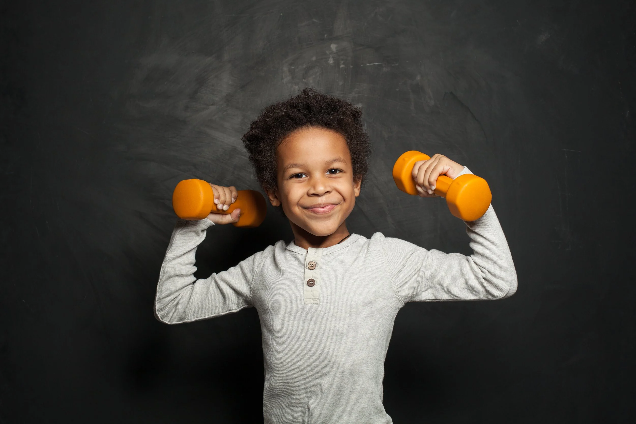 Smiling young boy with curly hair lifting orange dumbbells against a black chalkboard background.
