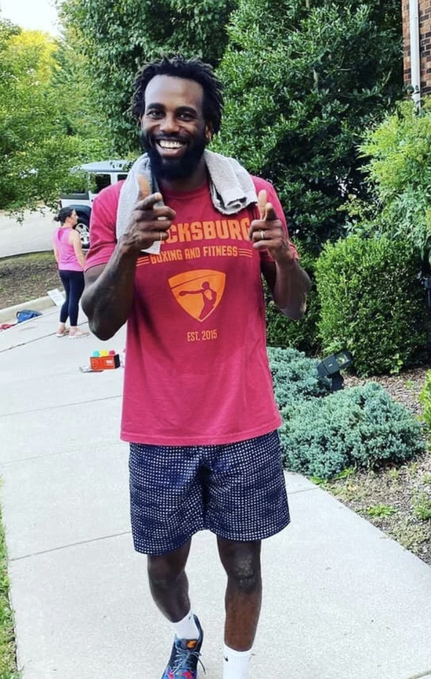 A man jogging outside, smiling and giving two thumbs up, wearing a red "Bucksburg Boxing and Fitness" t-shirt, black and white shorts, and sneakers, with a towel draped over his shoulders; background includes greenery and a woman in pink.