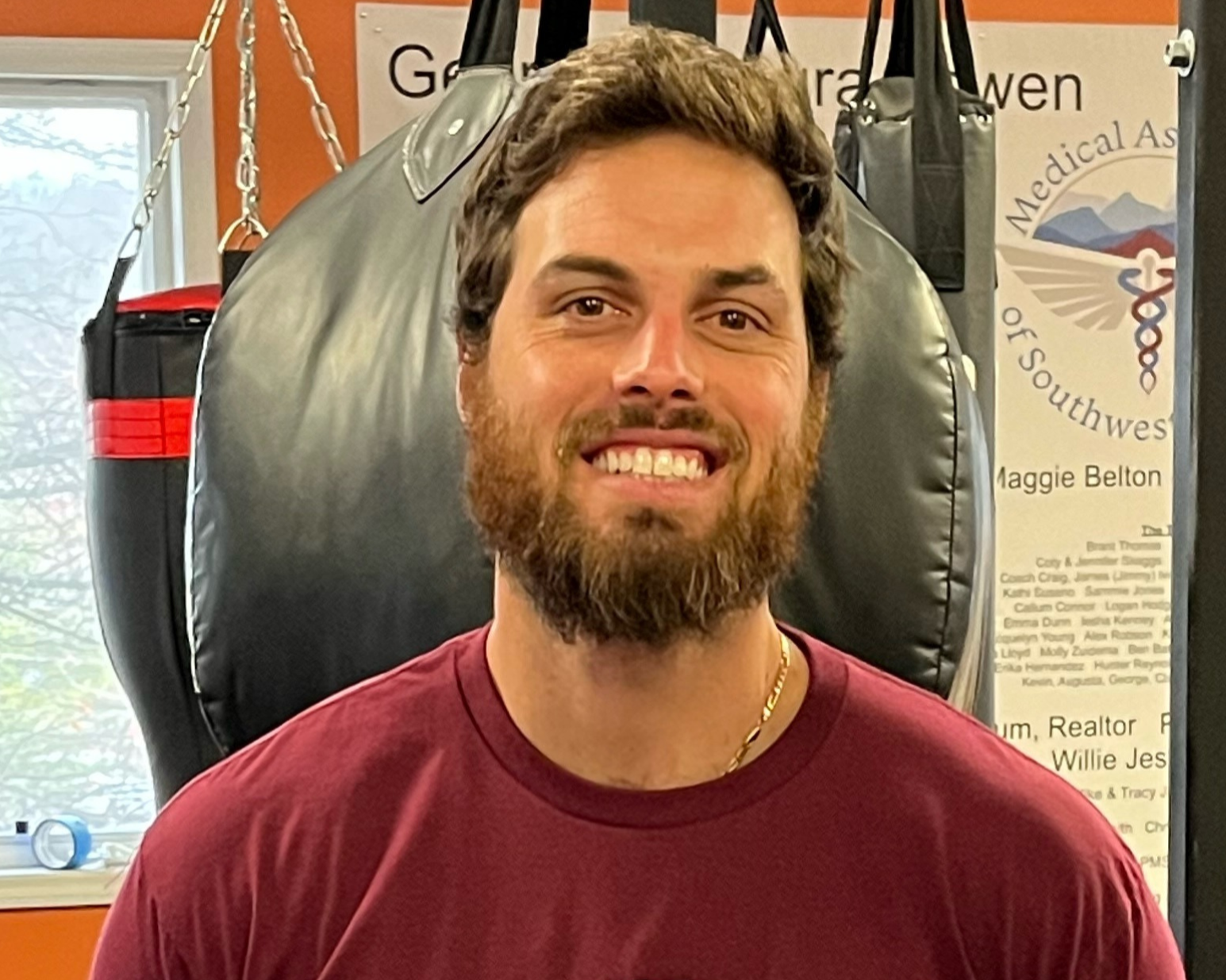 A young man with a beard and mustache smiling, wearing a maroon shirt and a gold chain, standing in front of punching bags and a sign about the Medical As...