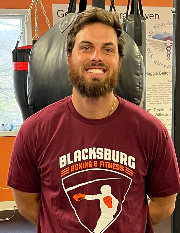 A man with a beard and curly hair smiling, wearing a maroon t-shirt with 'Blacksburg Boxing & Fitness' and a graphic of a person boxing. He is standing in a gym with a black punching bag behind him.