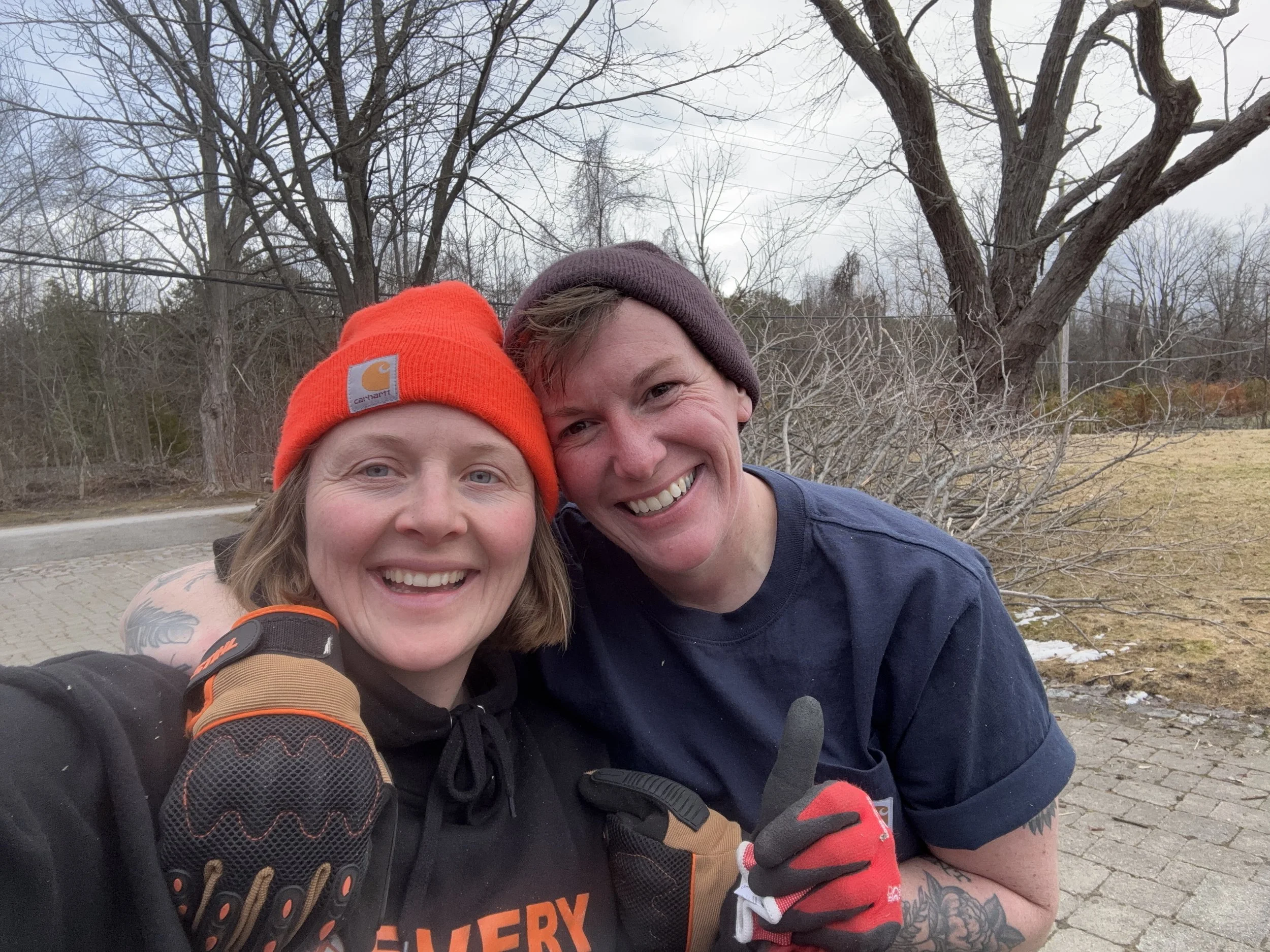 Two smiling women (Summerlong Garden owners Courtney-Anne and Jenna) taking a selfie outdoors on a cloudy day, wearing toques and gloves, with leafless trees and a paved path in the background.
