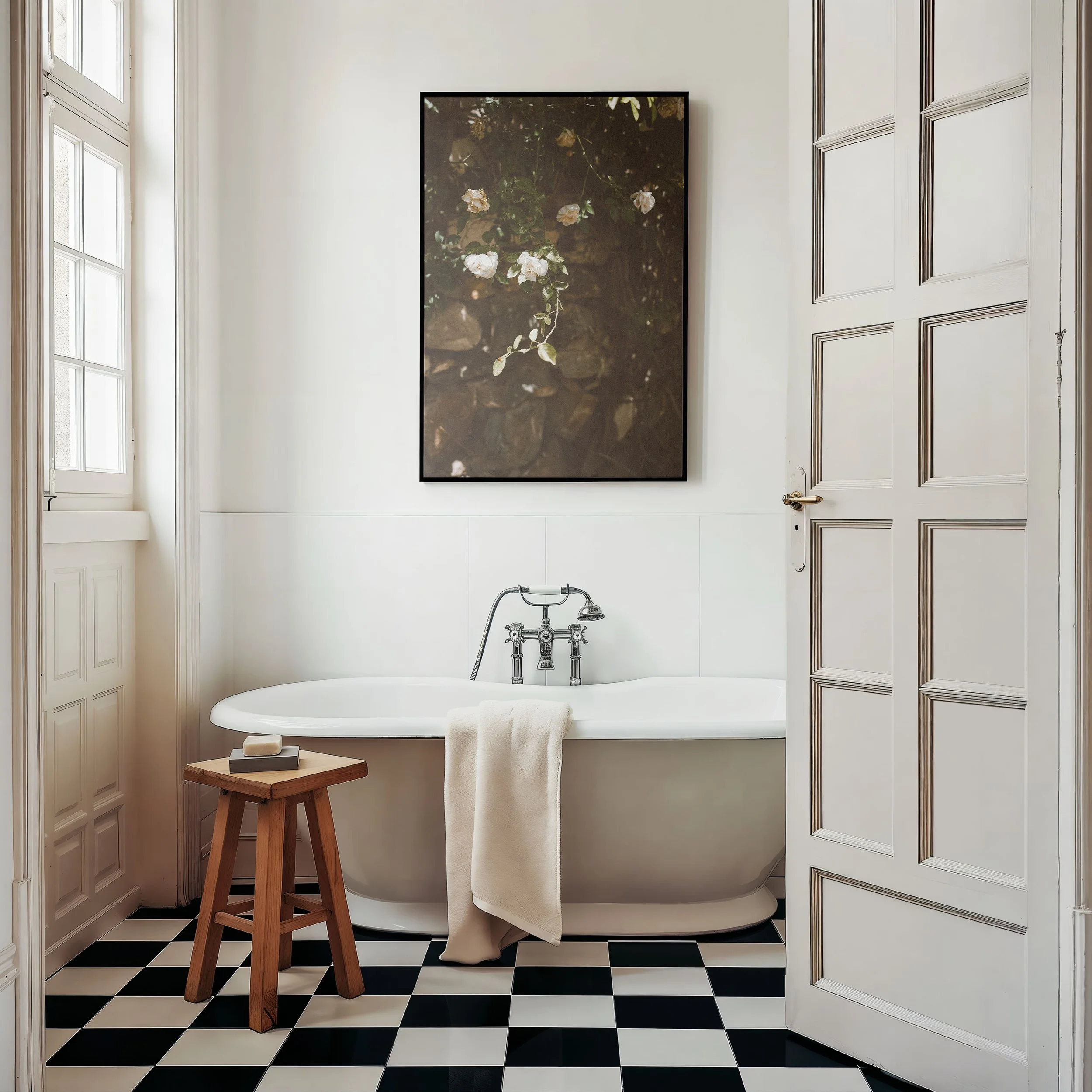 A white bathtub with a vintage silver faucet, a white towel hanging over the side, a small wooden stool with a soap and tray on top, a black and white checkerboard floor, a large framed artwork with botanical print on the wall, a window on the left, and an open white paneled door on the right.