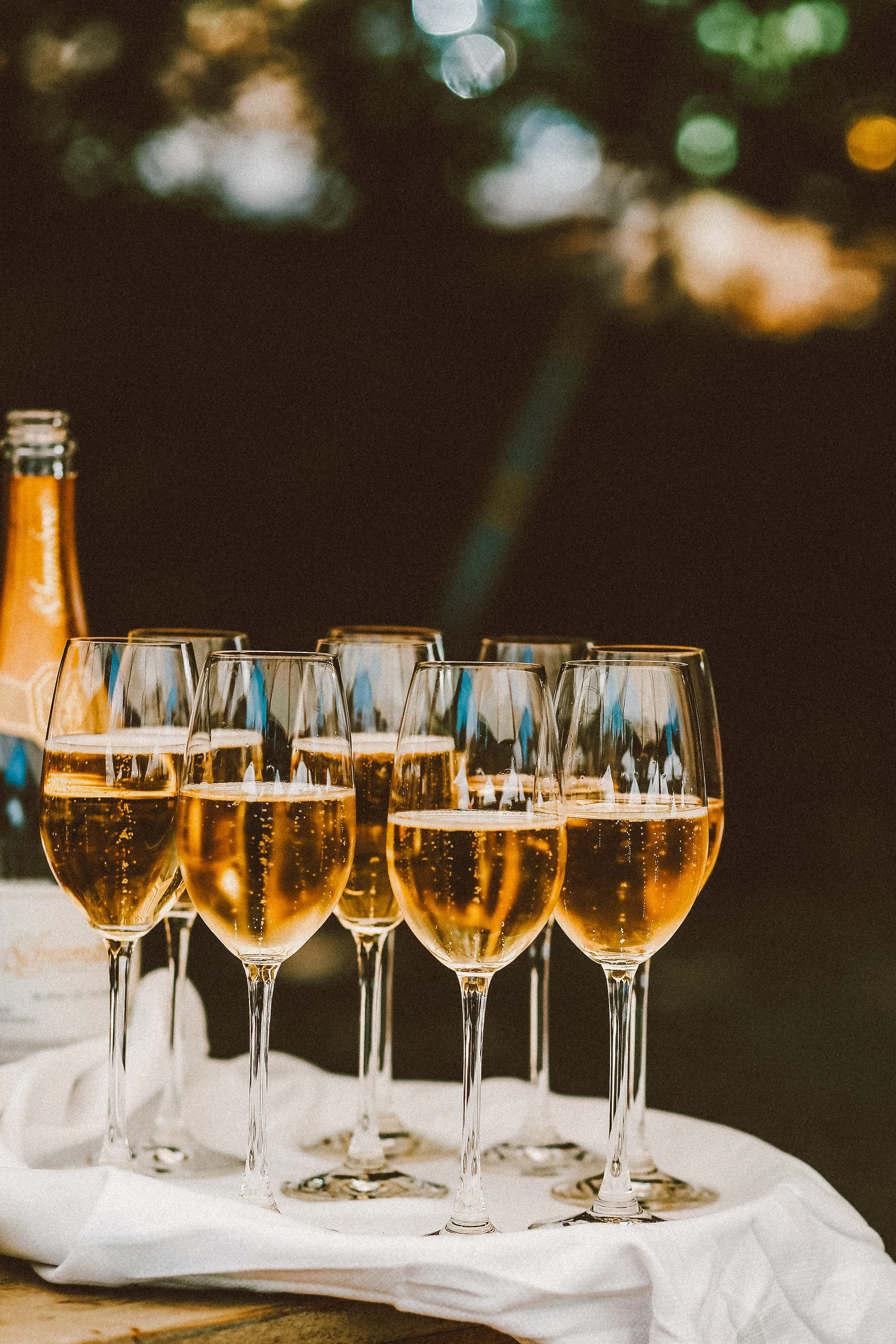 Six glasses of sparkling wine or champagne on a white cloth, with a blurred outdoor background.