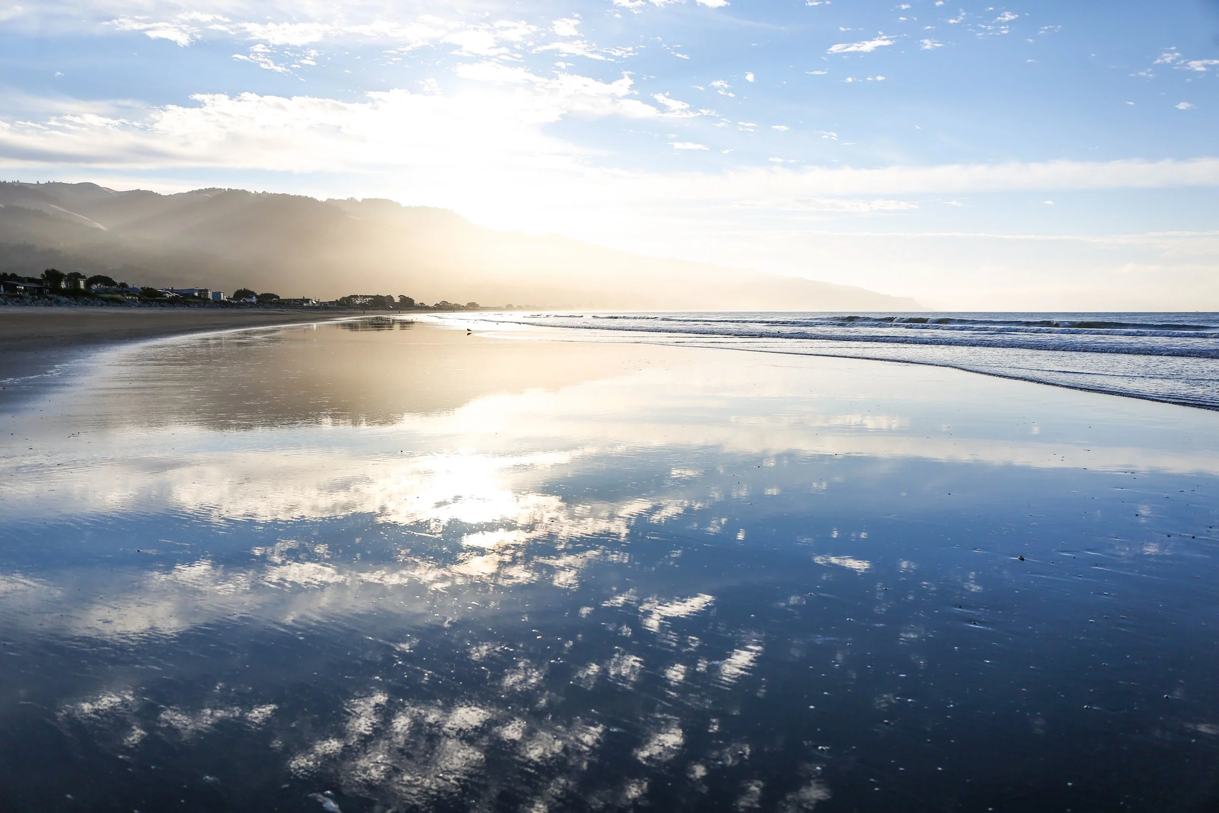 Sunlit beach with calm waves, reflecting clouds and sky, hills in the background.