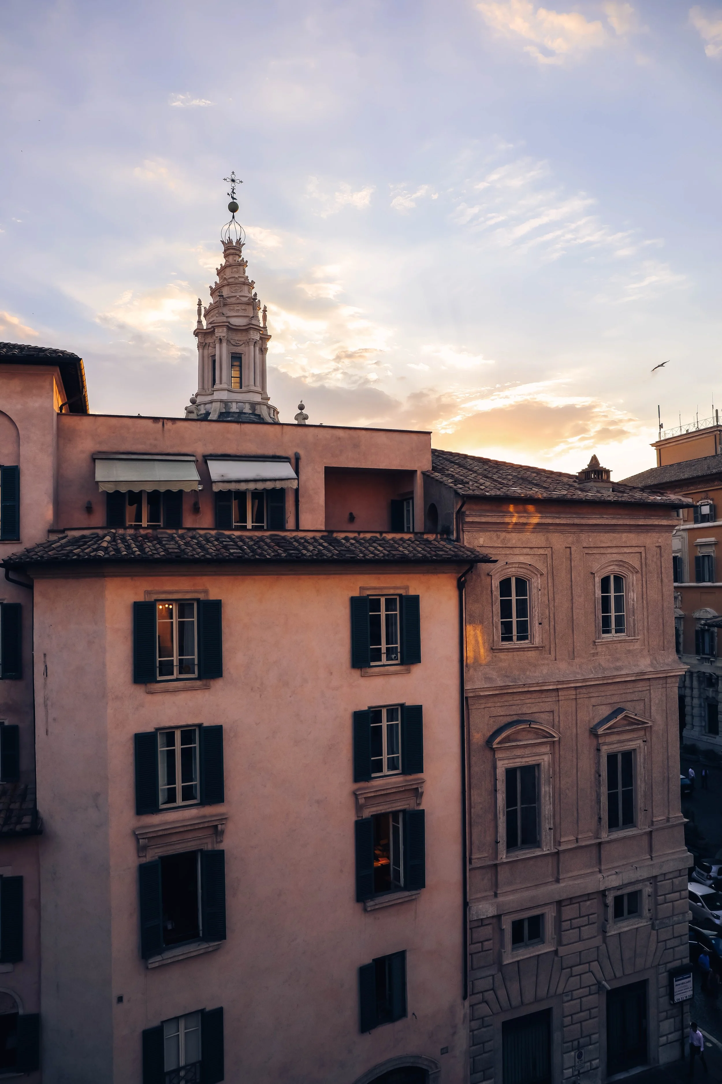 Sunset over European-style buildings with a church tower in the background, featuring a decorative cross and dome, in a cityscape.
