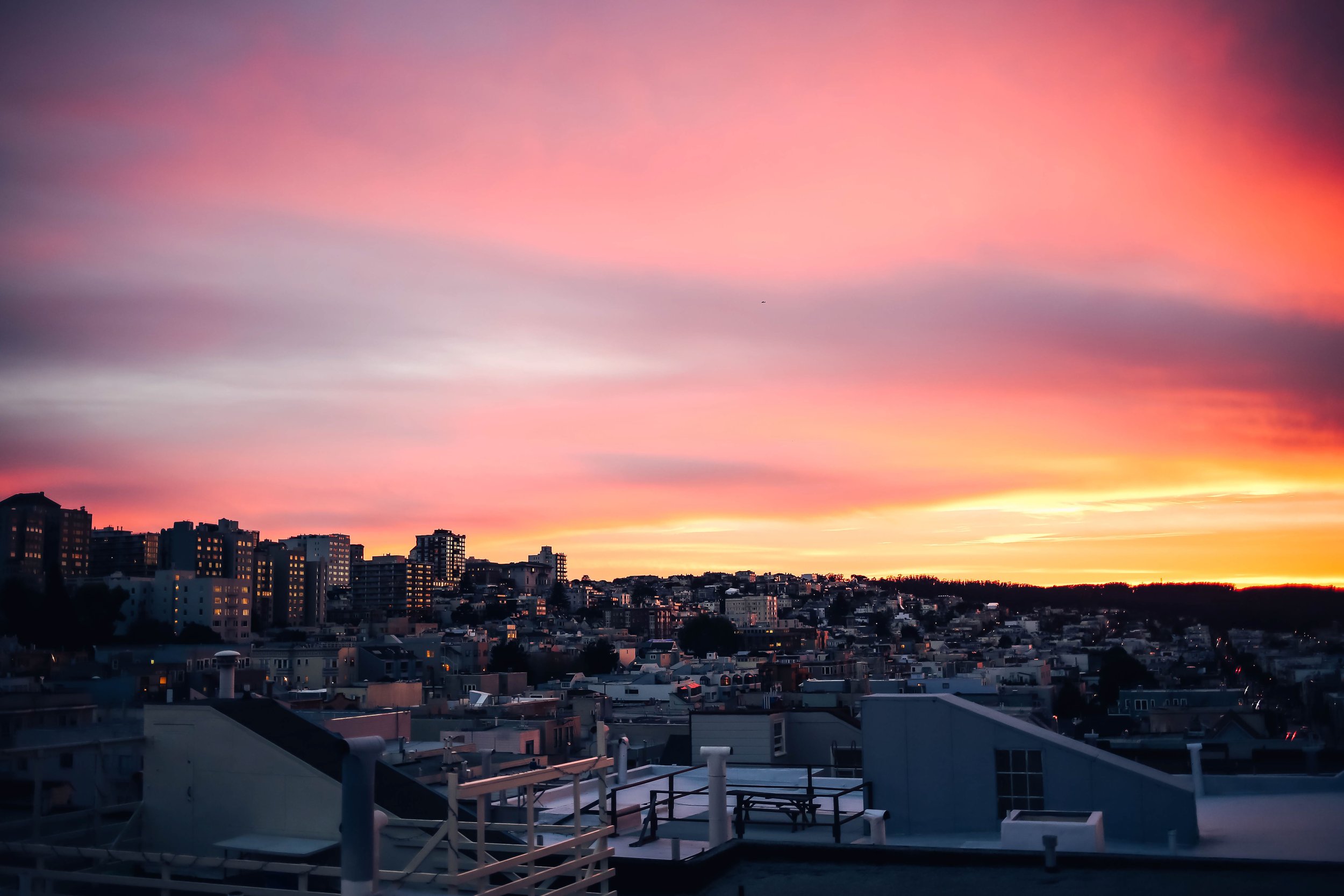 Pink and orange sunset sky over a hillside city skyline with lit windows and rooftops in the foreground