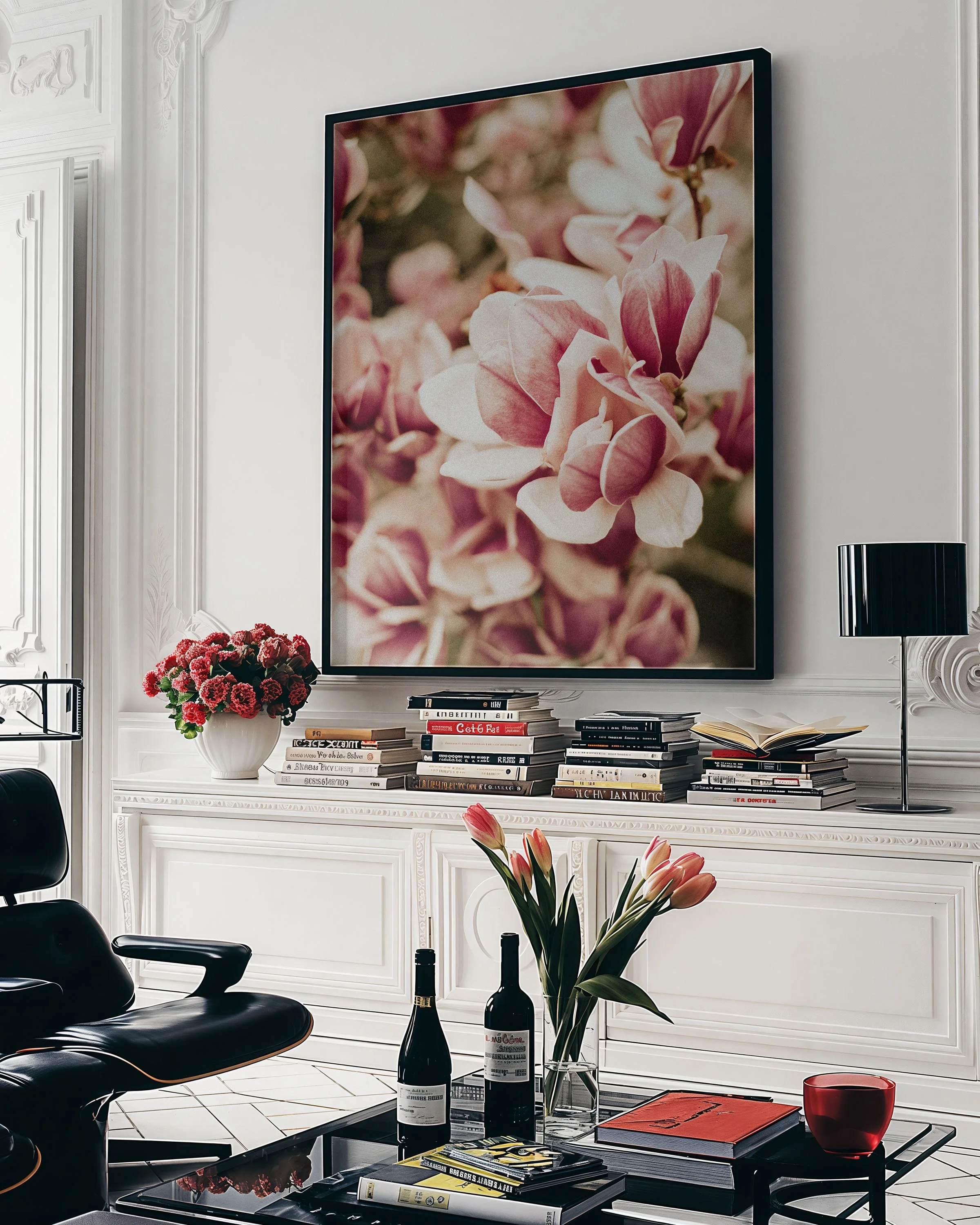 A living room with modern décor including a large framed floral artwork, a white console table with stacks of books, a potted flower, and a black chair. On the coffee table are wine bottles, a vase of pink tulips, and magazines.