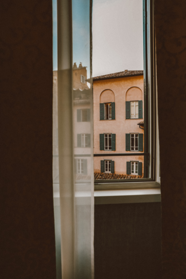 View through a partially open window showing a pink building with green shutters and a tiled roof.