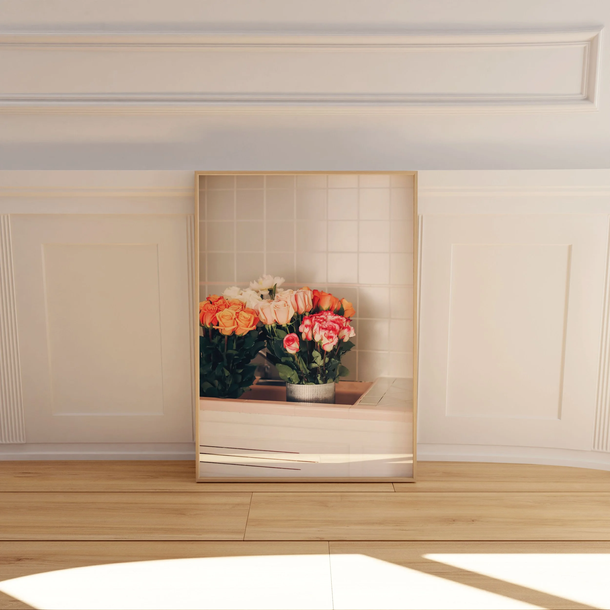 A framed mirror leaning against a white wainscoting wall, reflecting a bouquet of pink, white, and peach roses in a vase, with wood flooring and sunlight casting shadows.