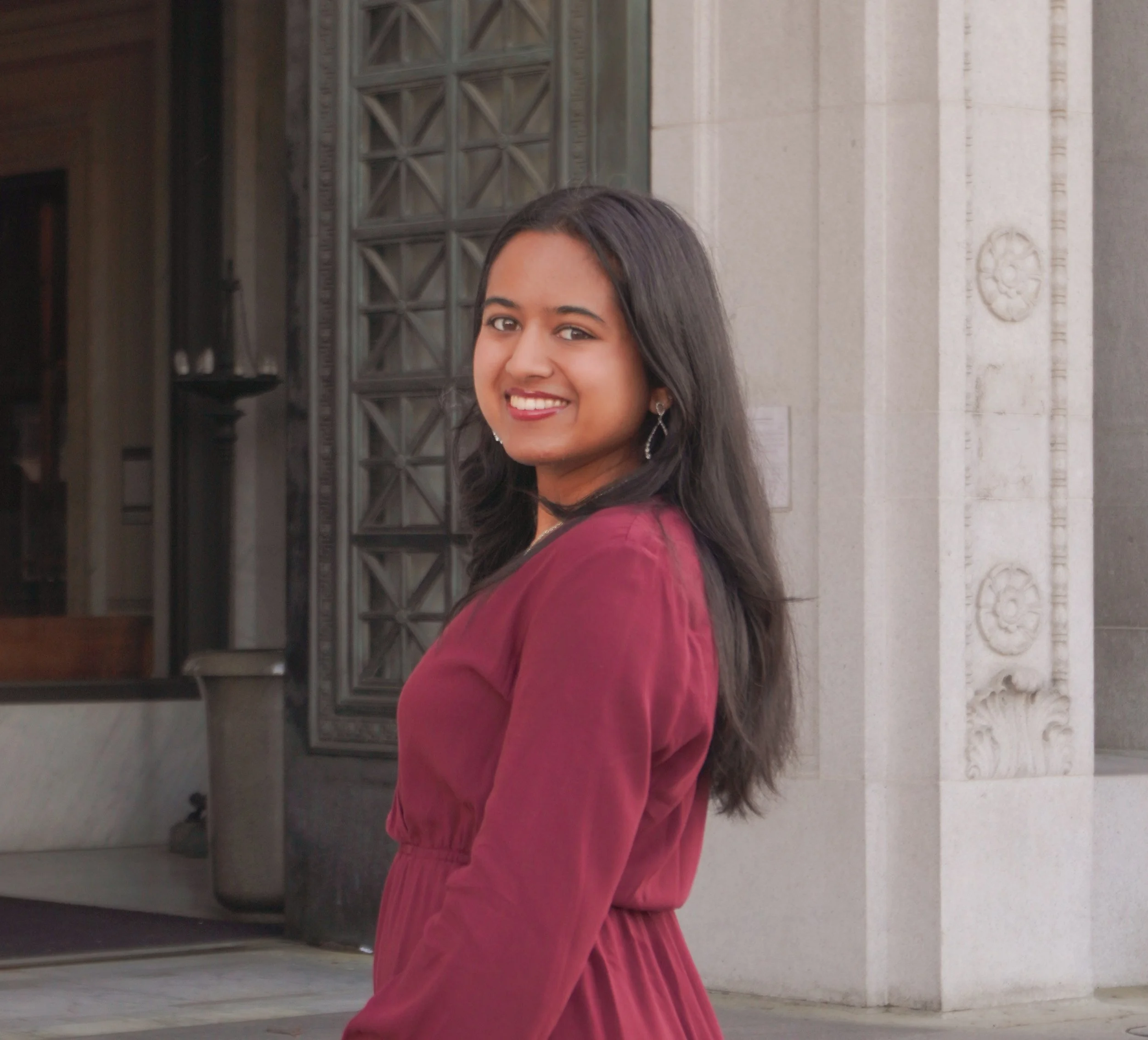 A smiling woman with long dark hair, wearing a maroon dress, standing outside a building with detailed stonework and large windows.