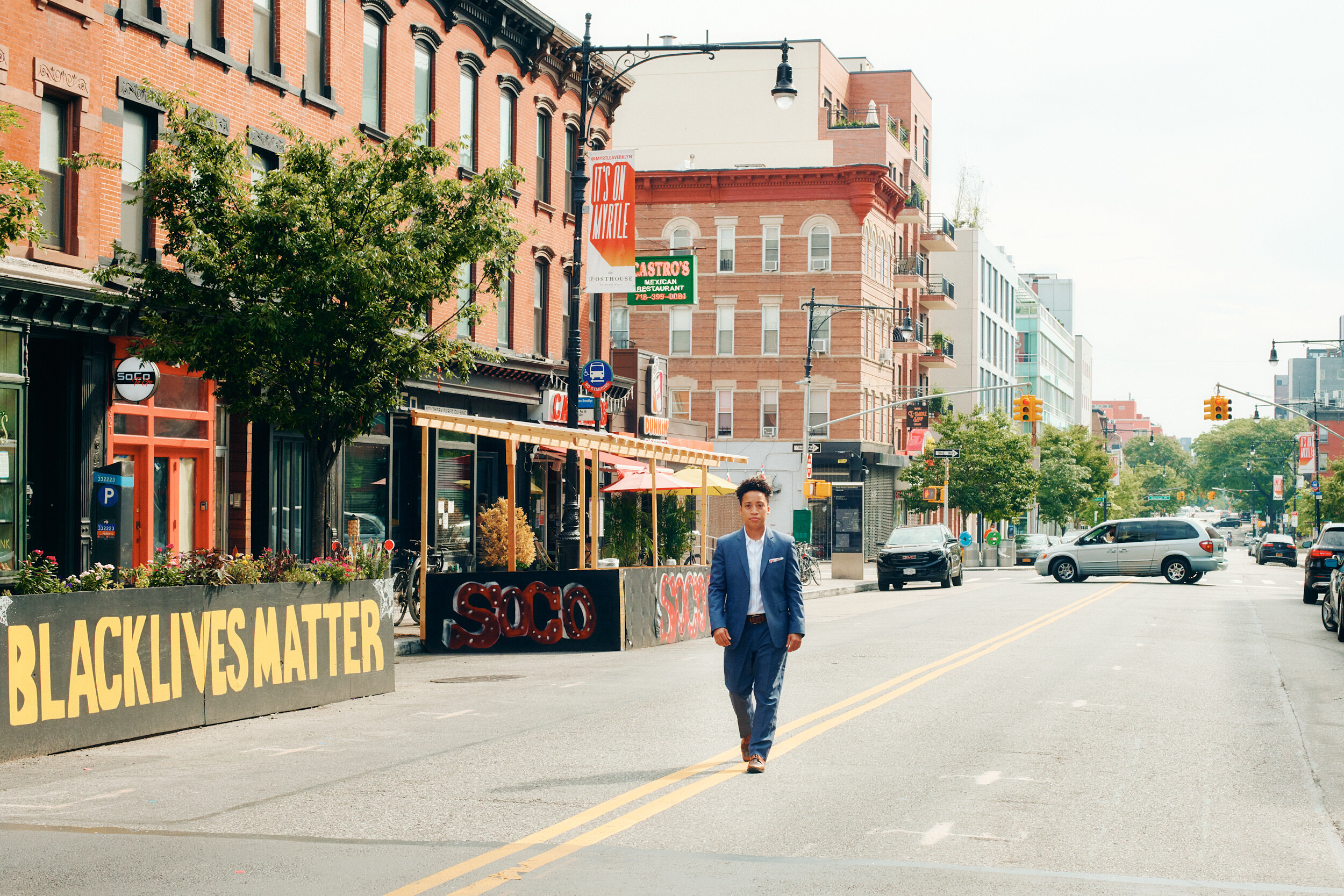 Far-away shot of Crystal walking down Myrtle Ave in Brooklyn wearing a Navy suit. In the background, there is a Black Lives Matter sign.