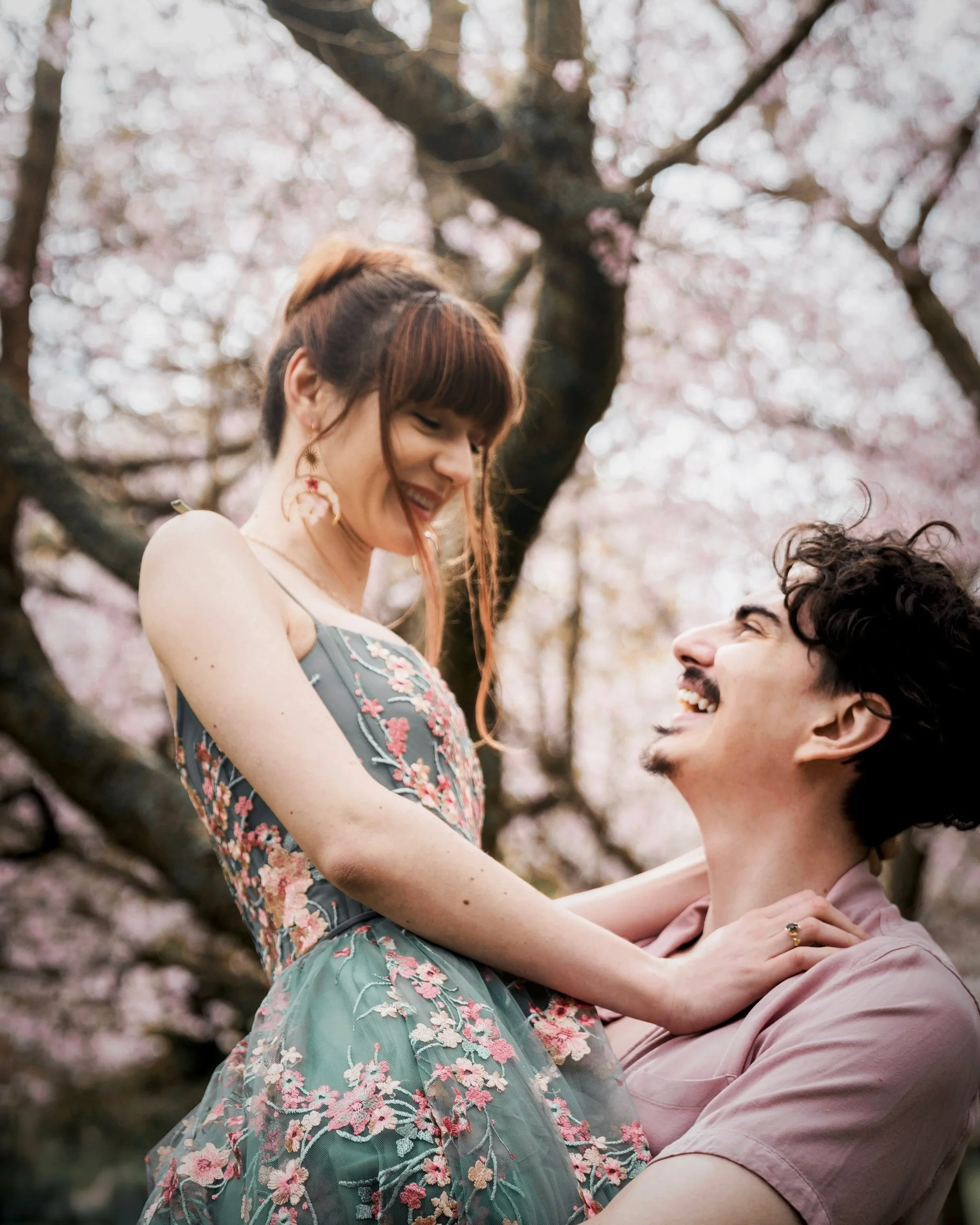 A smiling couple, woman wearing a floral dress and man in a pink shirt, enjoying a moment together among blooming cherry blossoms.