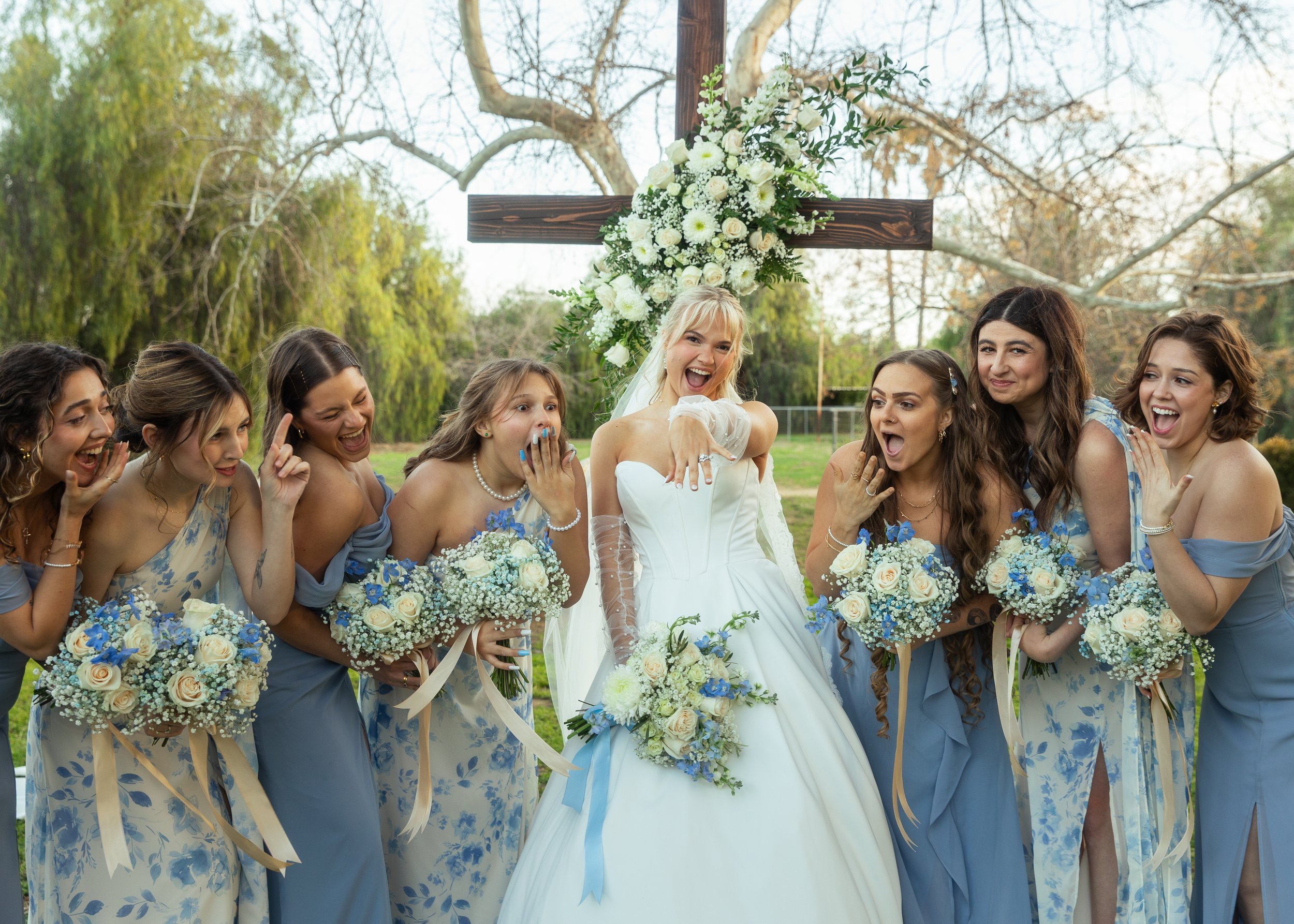 A bride in a white gown and her bridesmaids in light blue dresses smiling and posing outside in front of a large wooden cross decorated with white flowers.