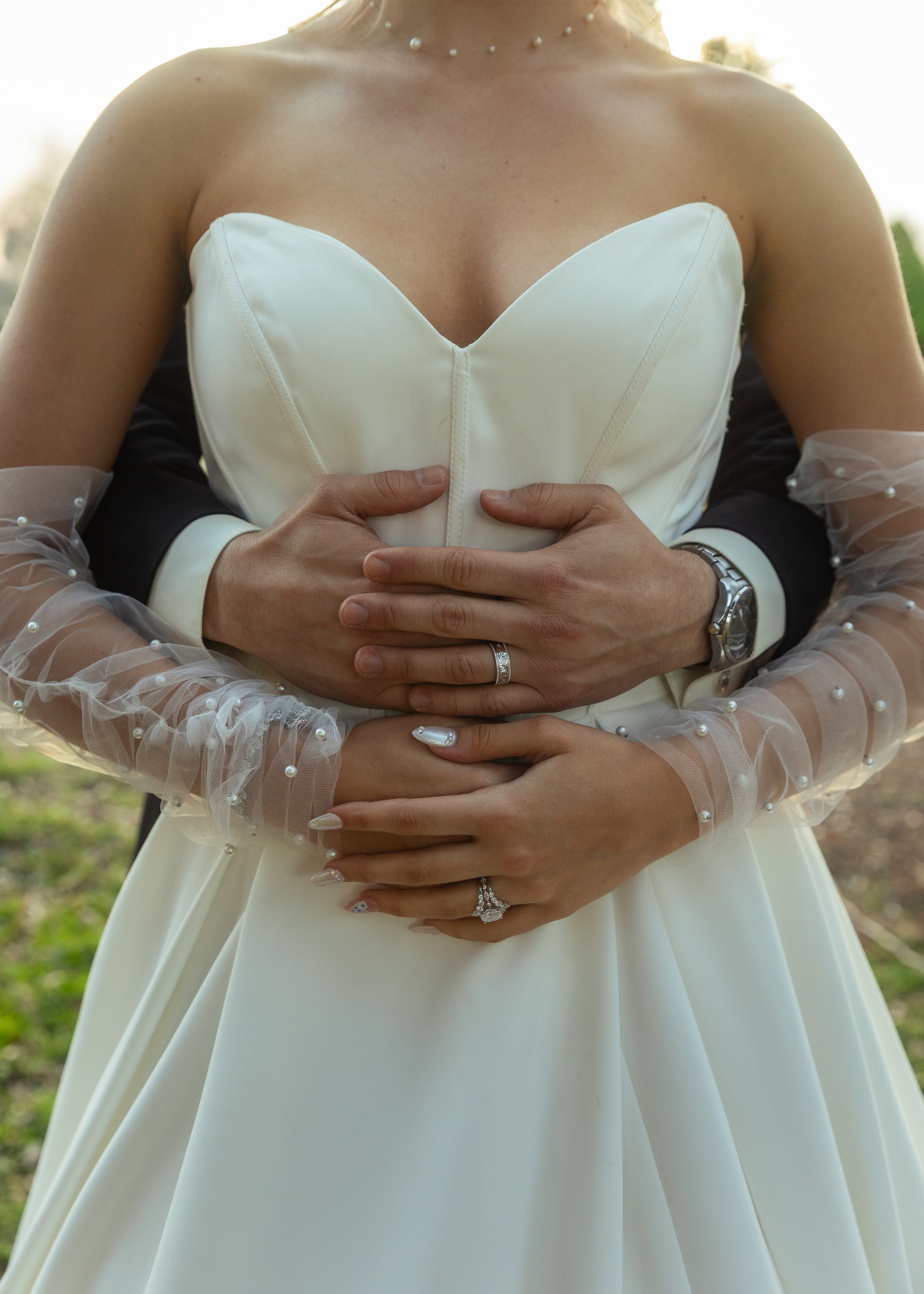 A bride in a white wedding dress with sheer, pearl-embellished sleeves, holding hands with someone behind her, both wearing watches and rings, outdoor setting with soft focus background.