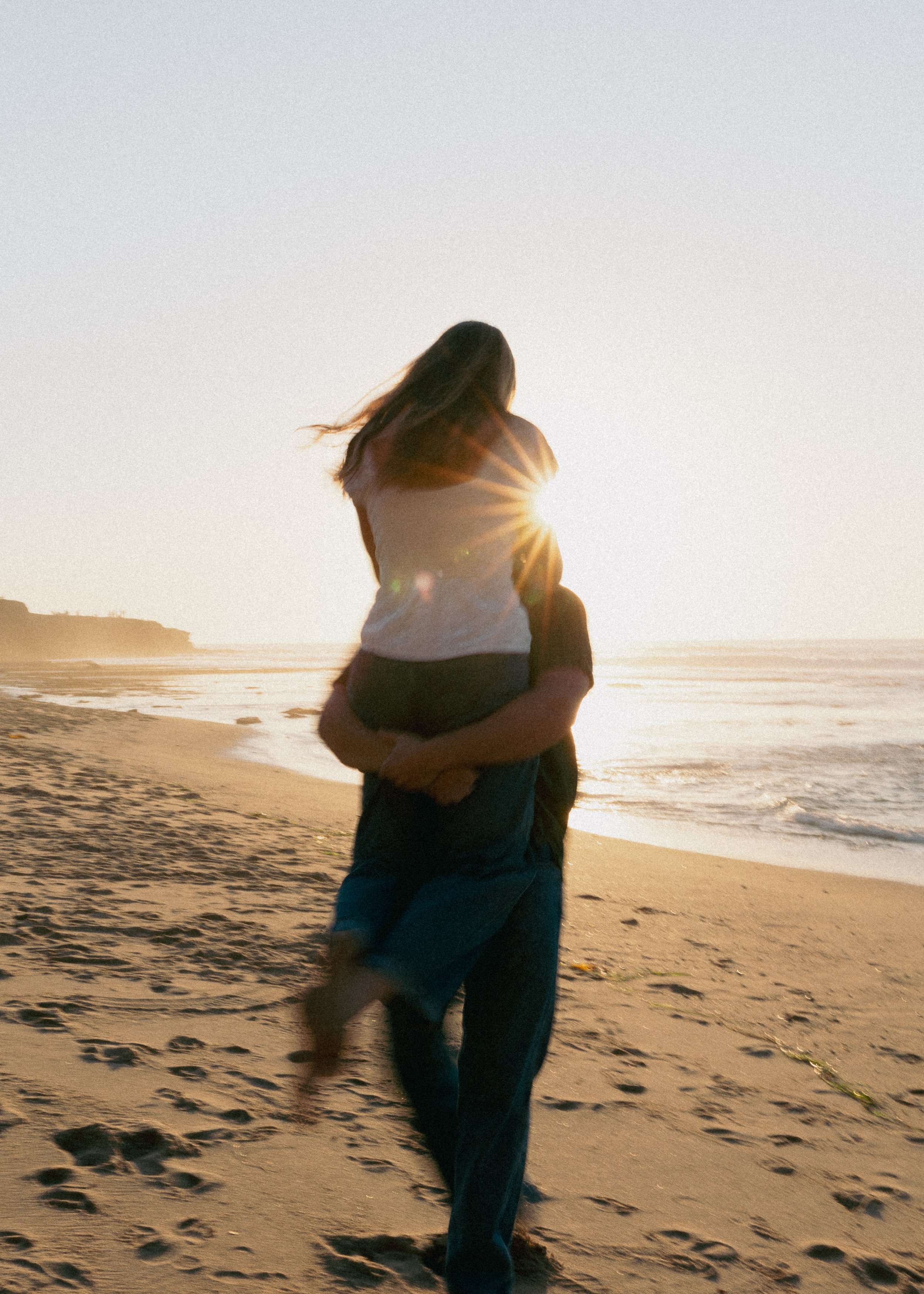 A couple on the beach at sunset, with one person sitting on the other's shoulders, as the sun creates a starburst effect behind them.