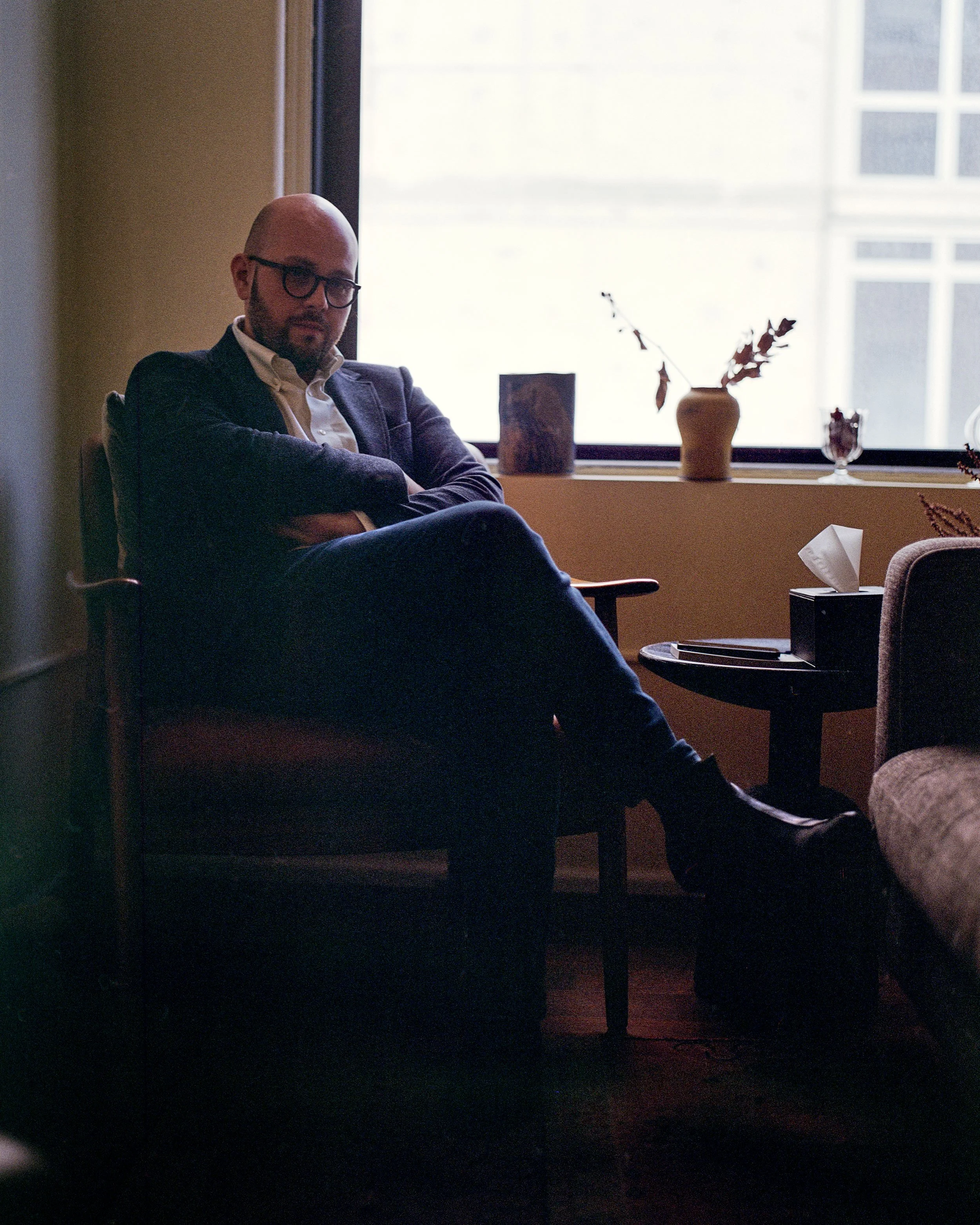 A man with glasses, a beard, and dressed in a dark blazer, seated with arms crossed in a dimly lit room near a window with frosted glass, with decorative vases and a tissue box on the windowsill.