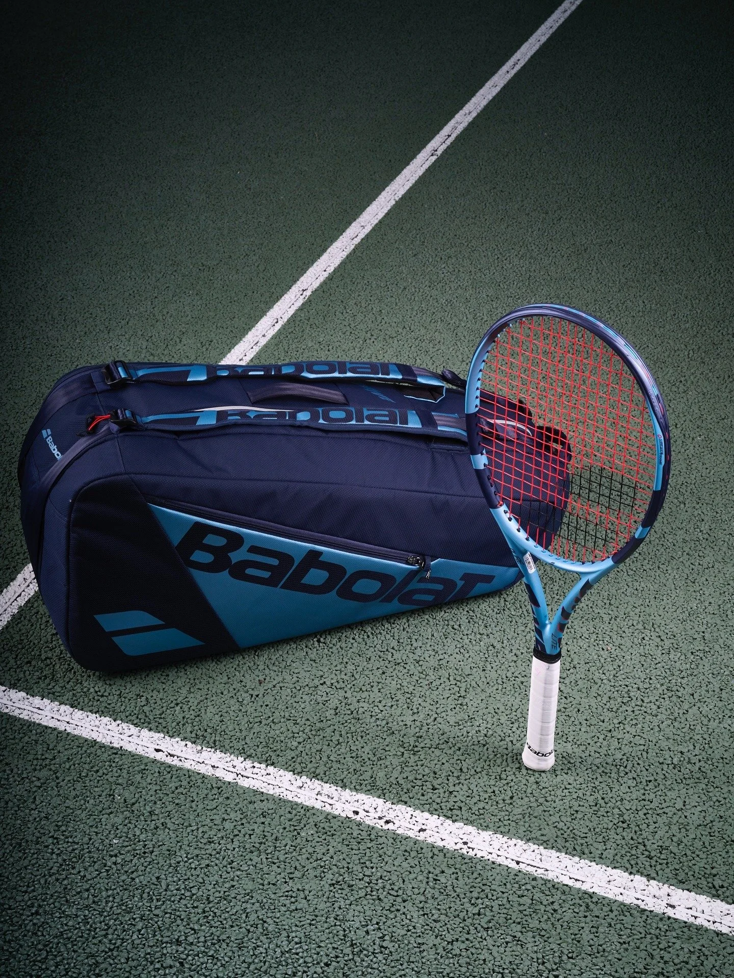 A Babolat tennis racket stands independently beside a blue Babolat tennis bag on a green outdoor tennis court with white lines.