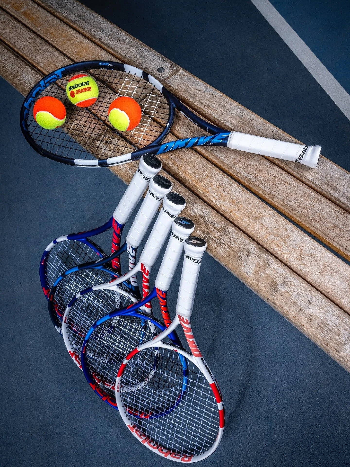 Five tennis racquets with white grips lean against a wooden bench on a tennis court. One racquet sits on top of the bench, with three yellow and orange tennis balls on it.