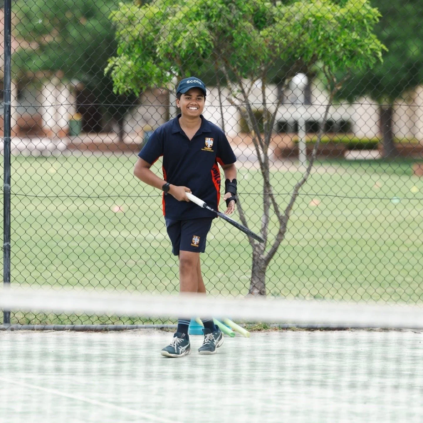 Smiling boy in a school sports uniform standing on a tennis court holding a tennis racquet.