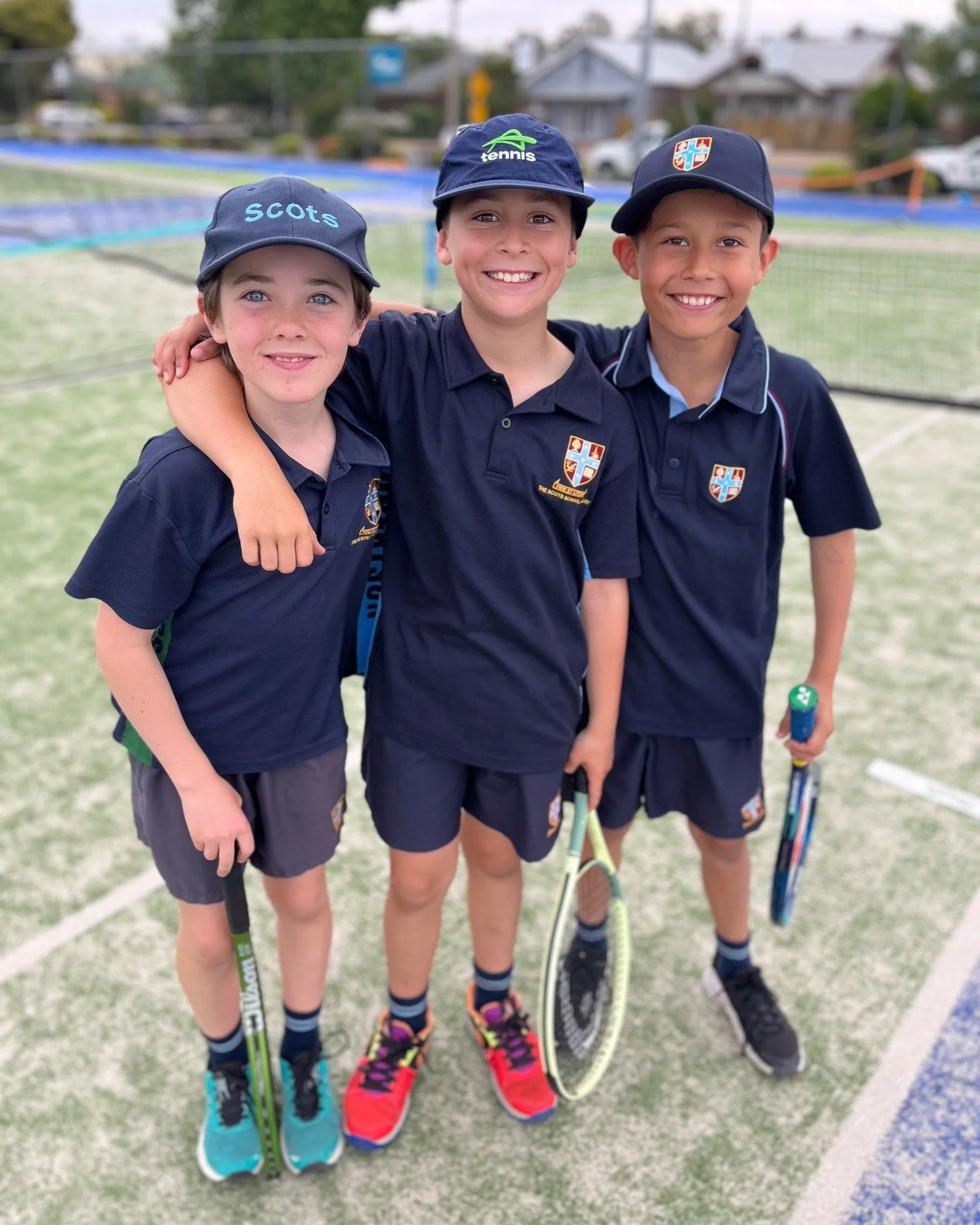 Three young boys in navy blue school uniforms standing on a tennis court, smiling, with tennis racquets in their hands, and their arms around each other.
