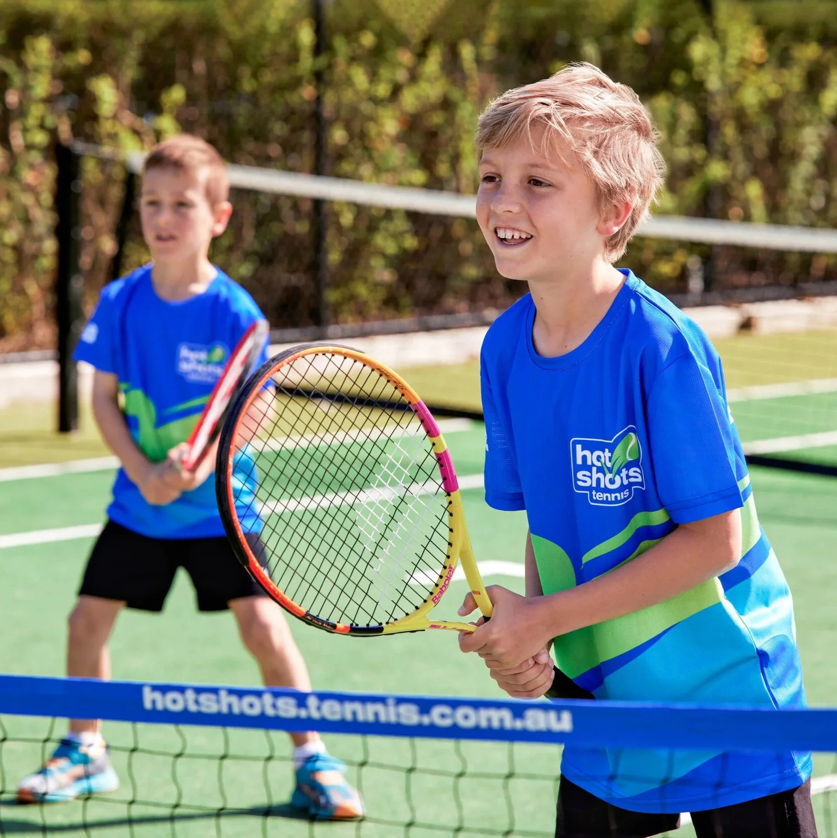Two young boys wearing blue Hot Shots uniforms stand at a tennis net with racquets in hand.