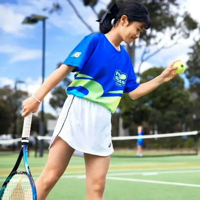A young girl in a blue Hot Shorts shirt and white tennis skirt holding a tennis ball in her left hand and a tennis racquet in her left hand on a tennis court.