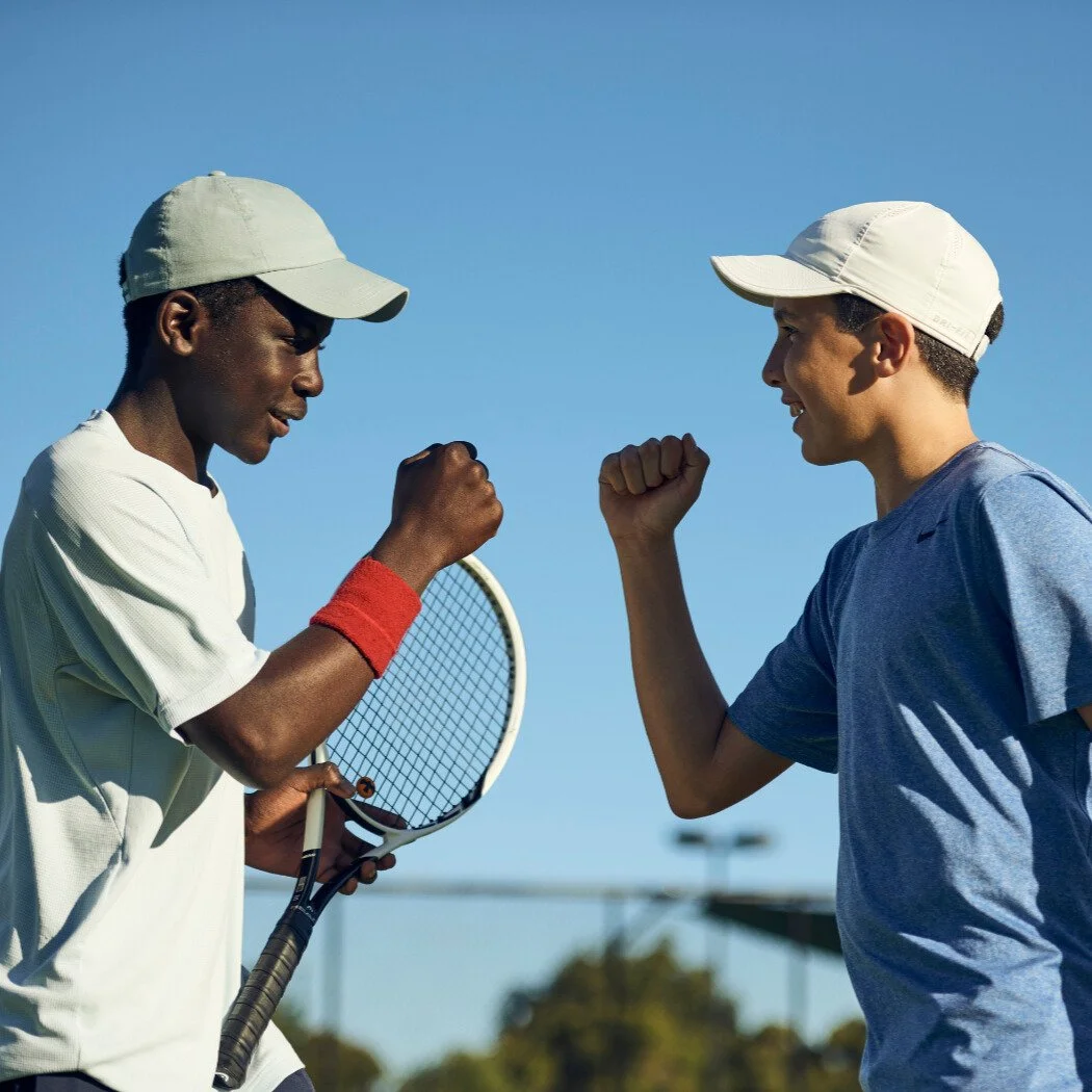 Two young tennis players fist-bumping on a court, holding racquets and smiling during a practice session.