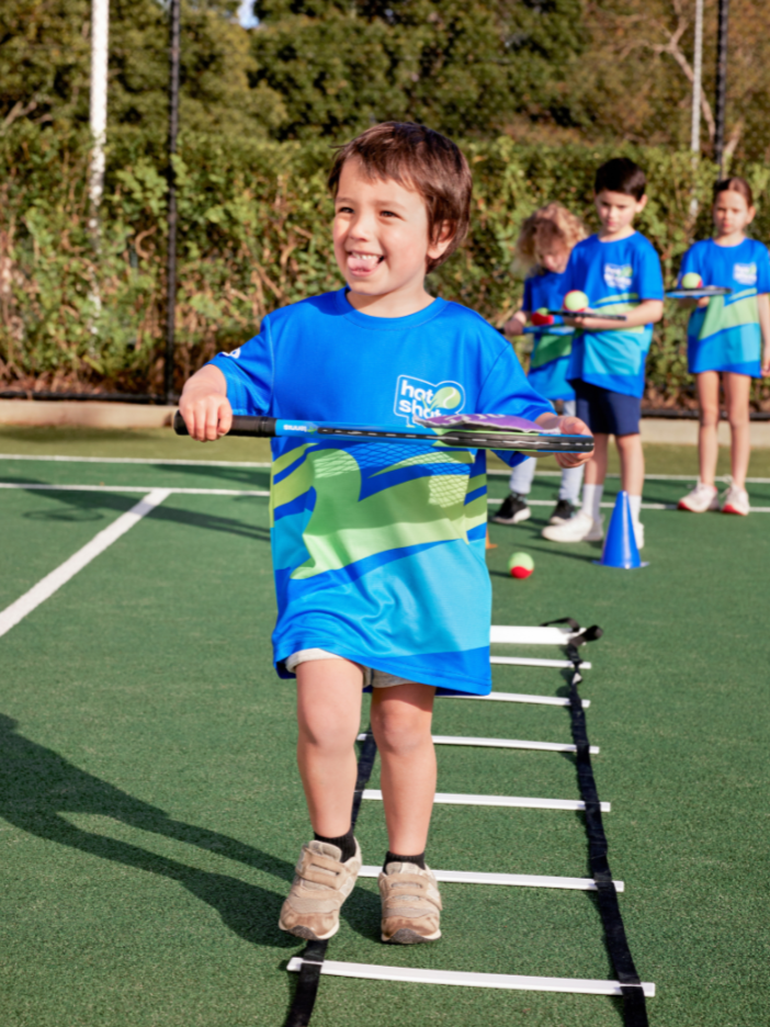 A young child in a Hot Shots tennis shirt smiling while stepping through an agility ladder during a tennis drill, with other children waiting in line behind.
