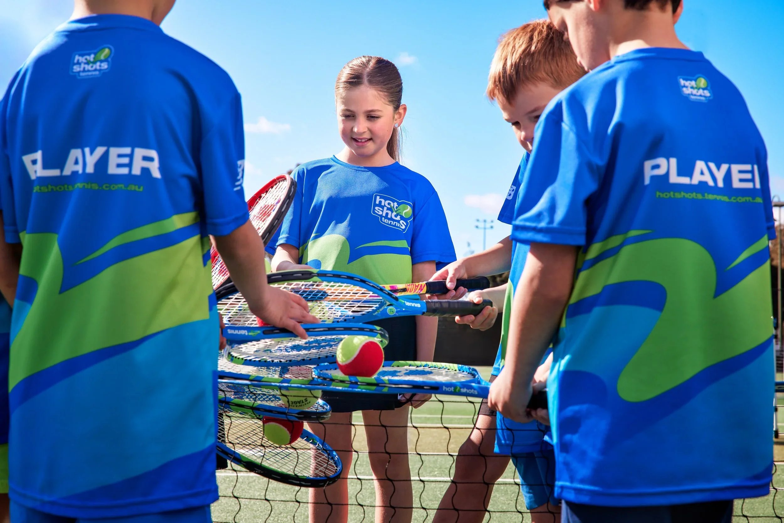 A group of children in Hot Shots tennis uniforms gathered at the net, holding colourful racquets and red tennis balls during a coaching session.