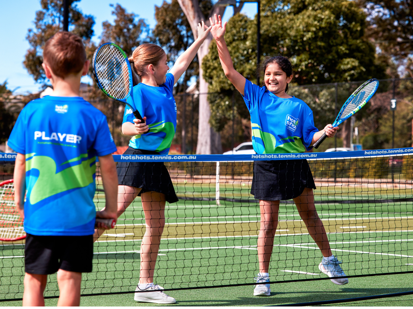 Two girls in Hot Shots tennis uniforms high-fiving over the net during a match on an outdoor court.