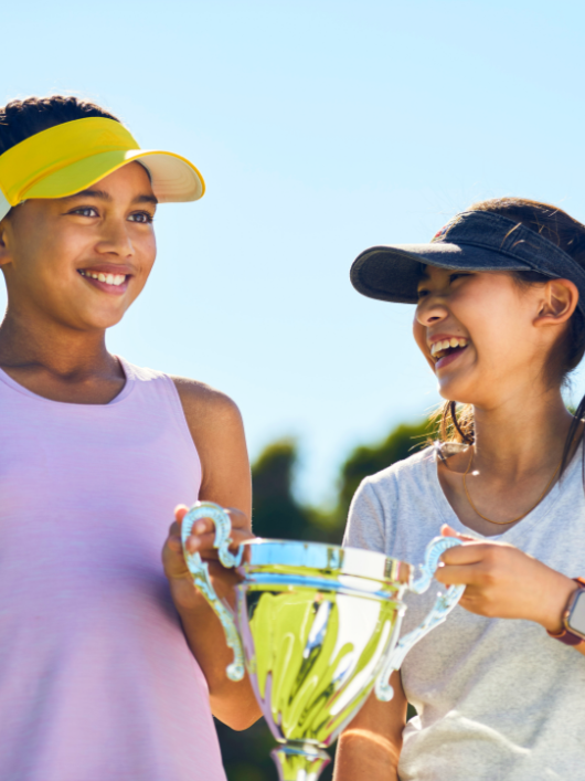 Two young female tennis players smiling and holding a trophy together on an outdoor court.